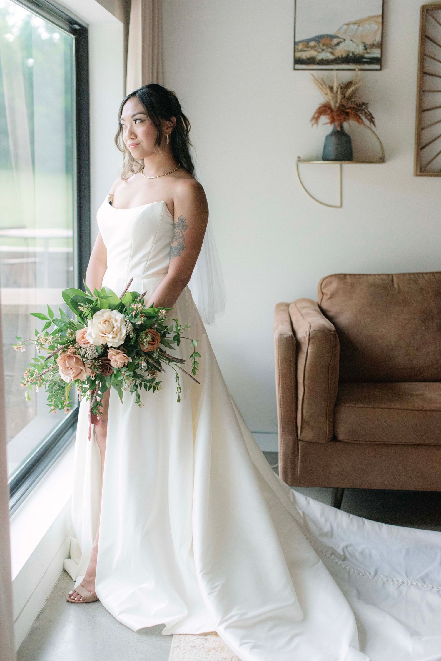 Bride in white wedding dress poses by window with floral bouquet in modern interior setting.