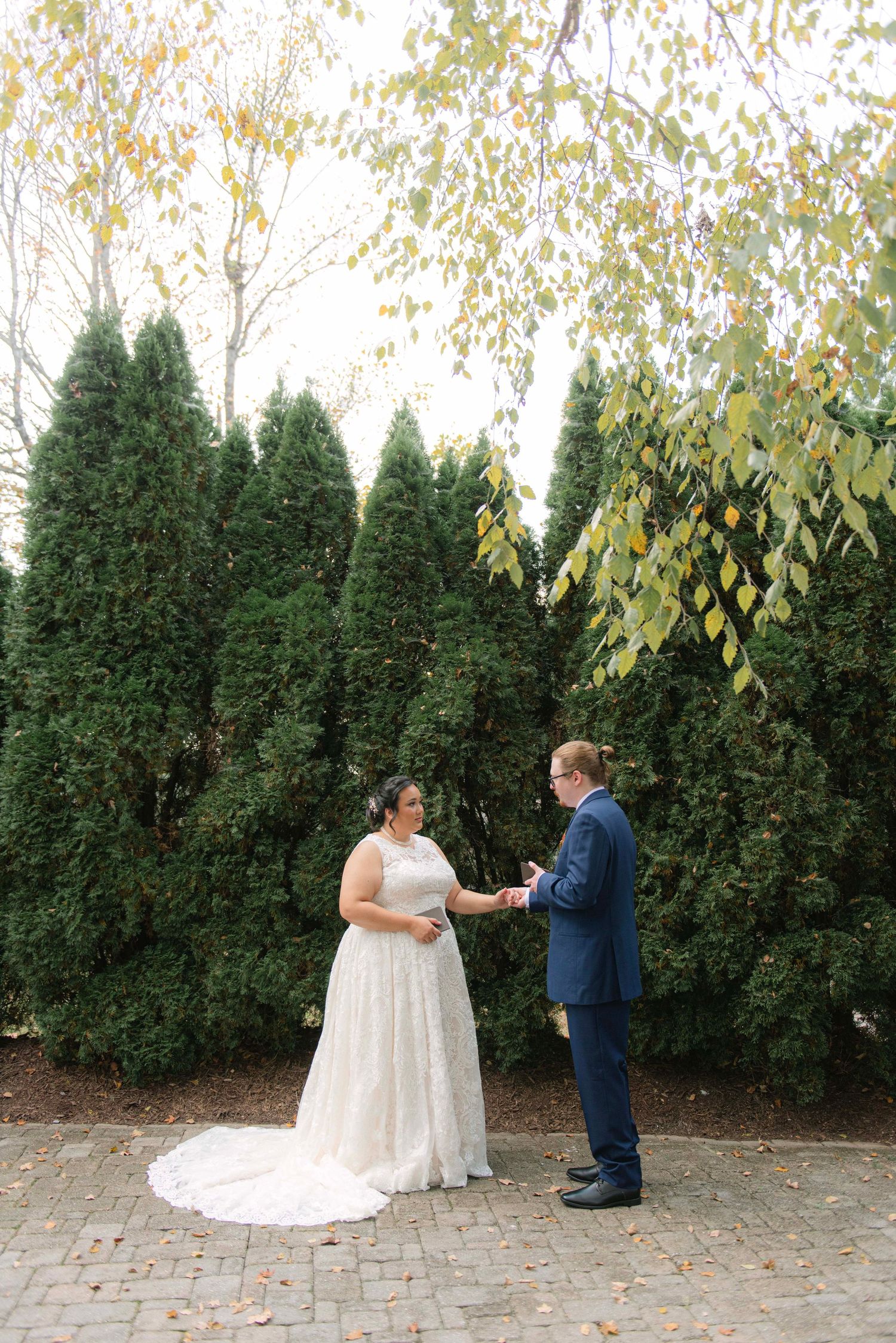Wedding couple exchanging vows in front of evergreen trees on a stone patio.
