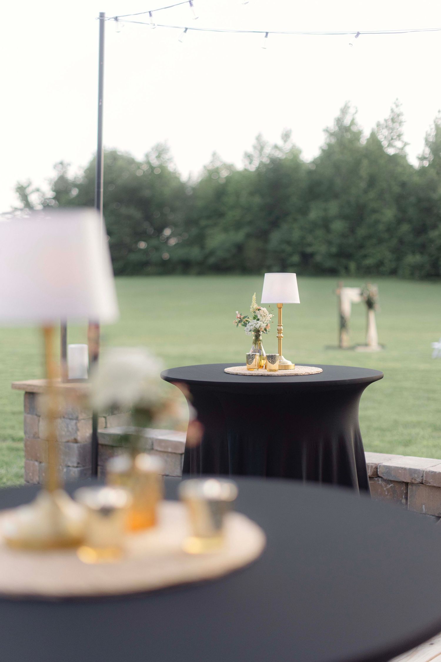 Elegant table setting with white linen lampshades and glassware arranged on a dark surface at an outdoor event.