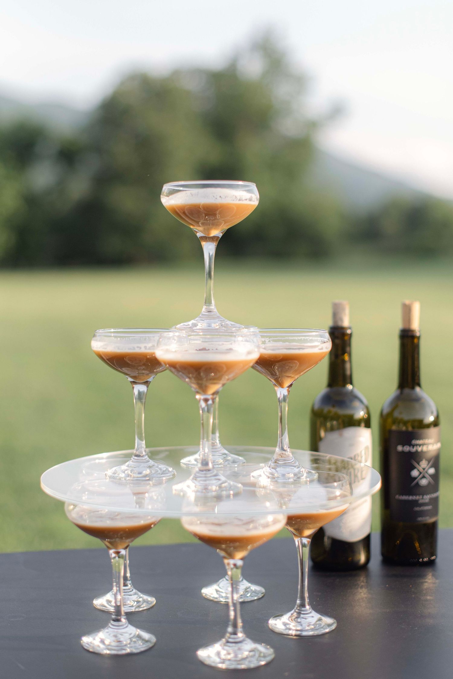 Elegant champagne coupe glasses filled with pink cocktails arranged on a dark table outdoors with wine bottles in background.