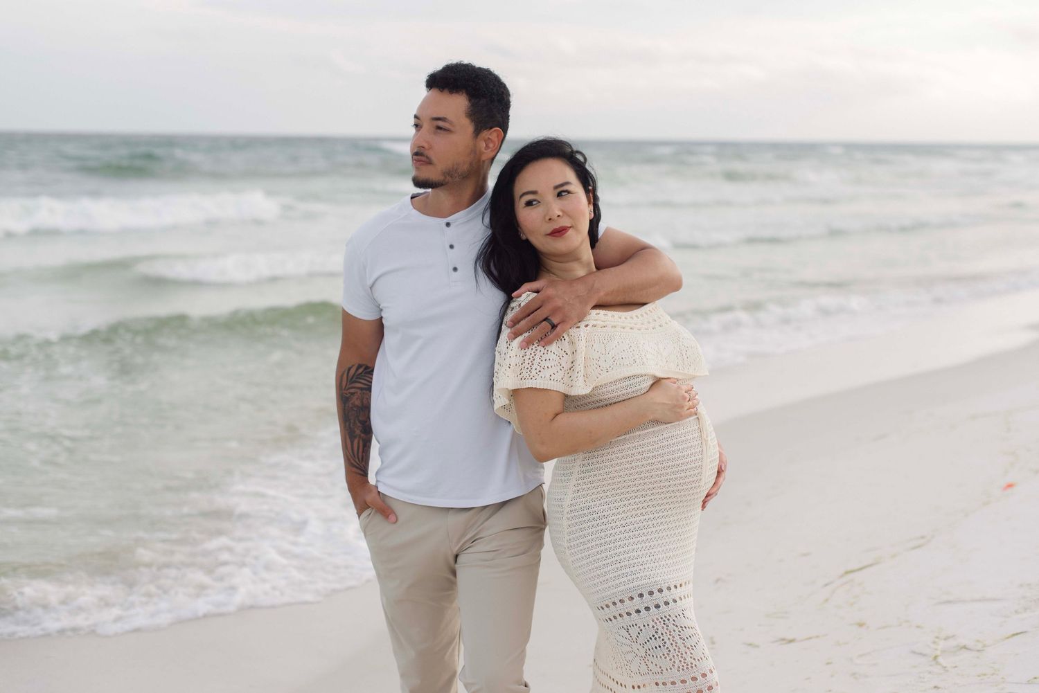 A couple in white and beige outfits dancing together on a scenic beach with waves in the background.