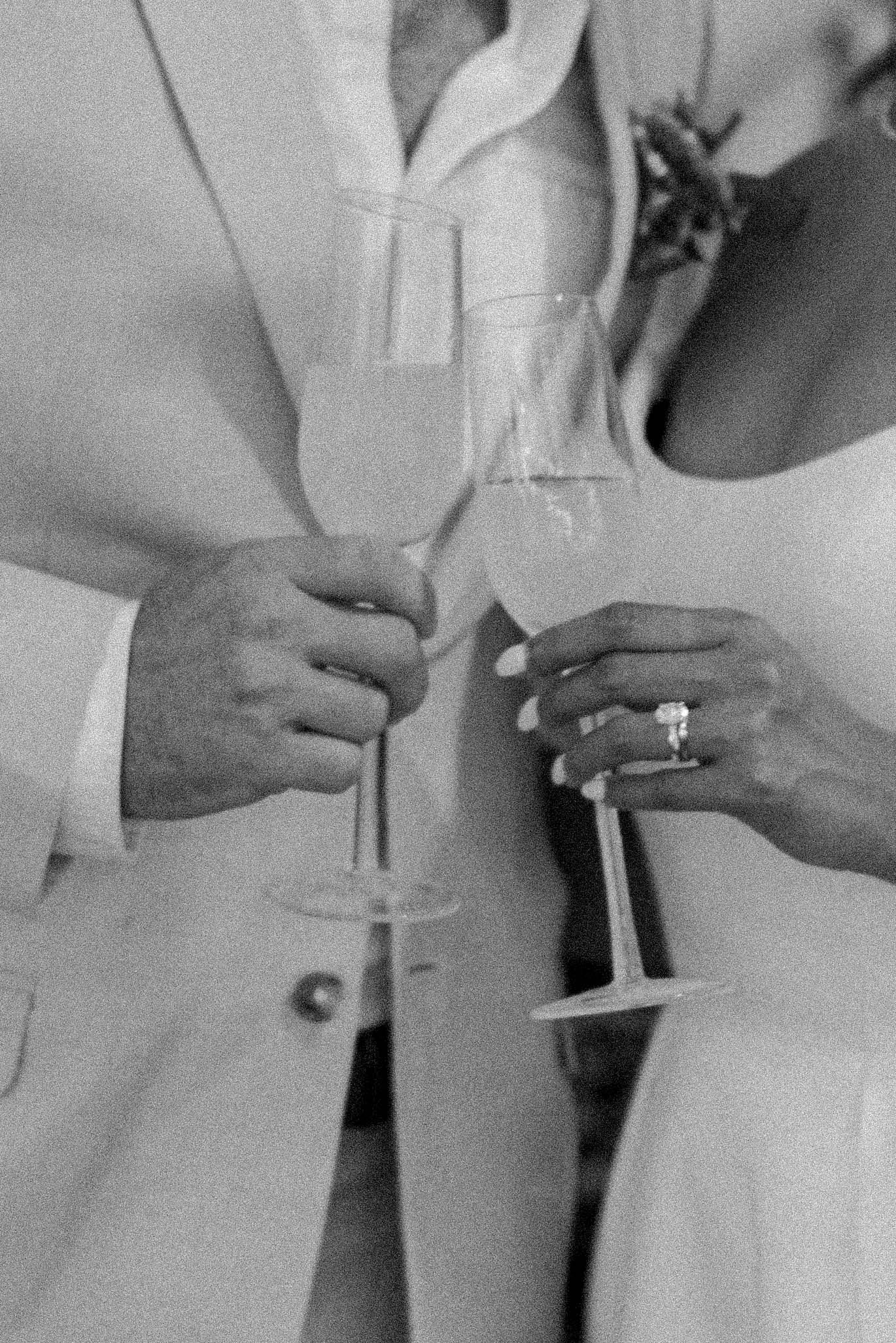 A black and white close-up of hands holding champagne glasses with a diamond engagement ring visible.