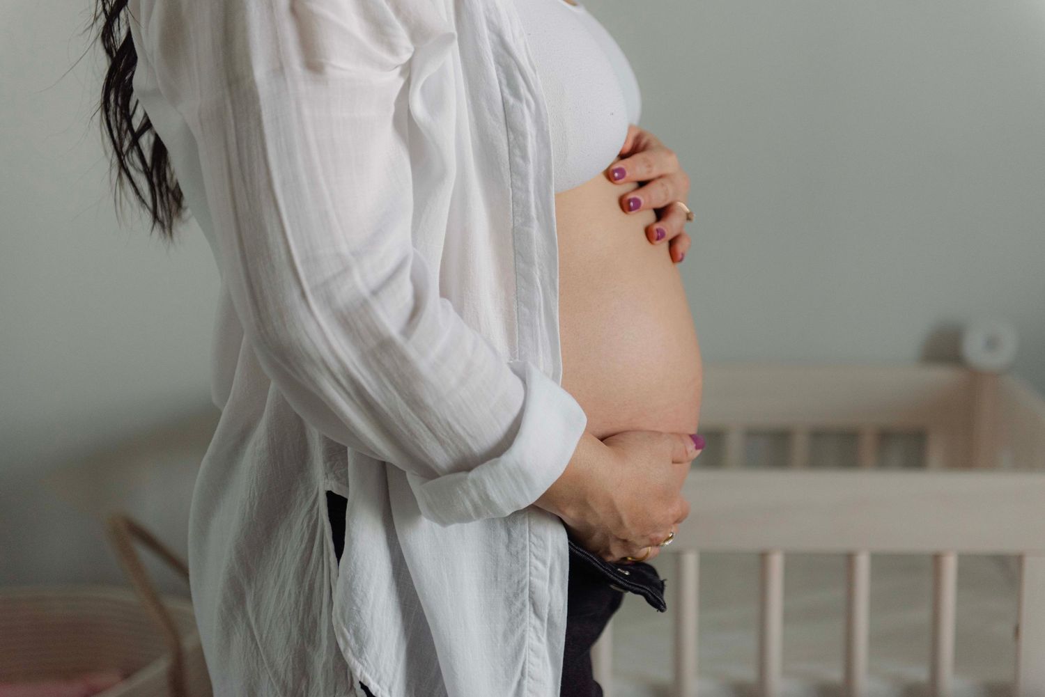 Side view of pregnant belly with white flowy shirt lifted to show bare bump, with wooden crib in background.