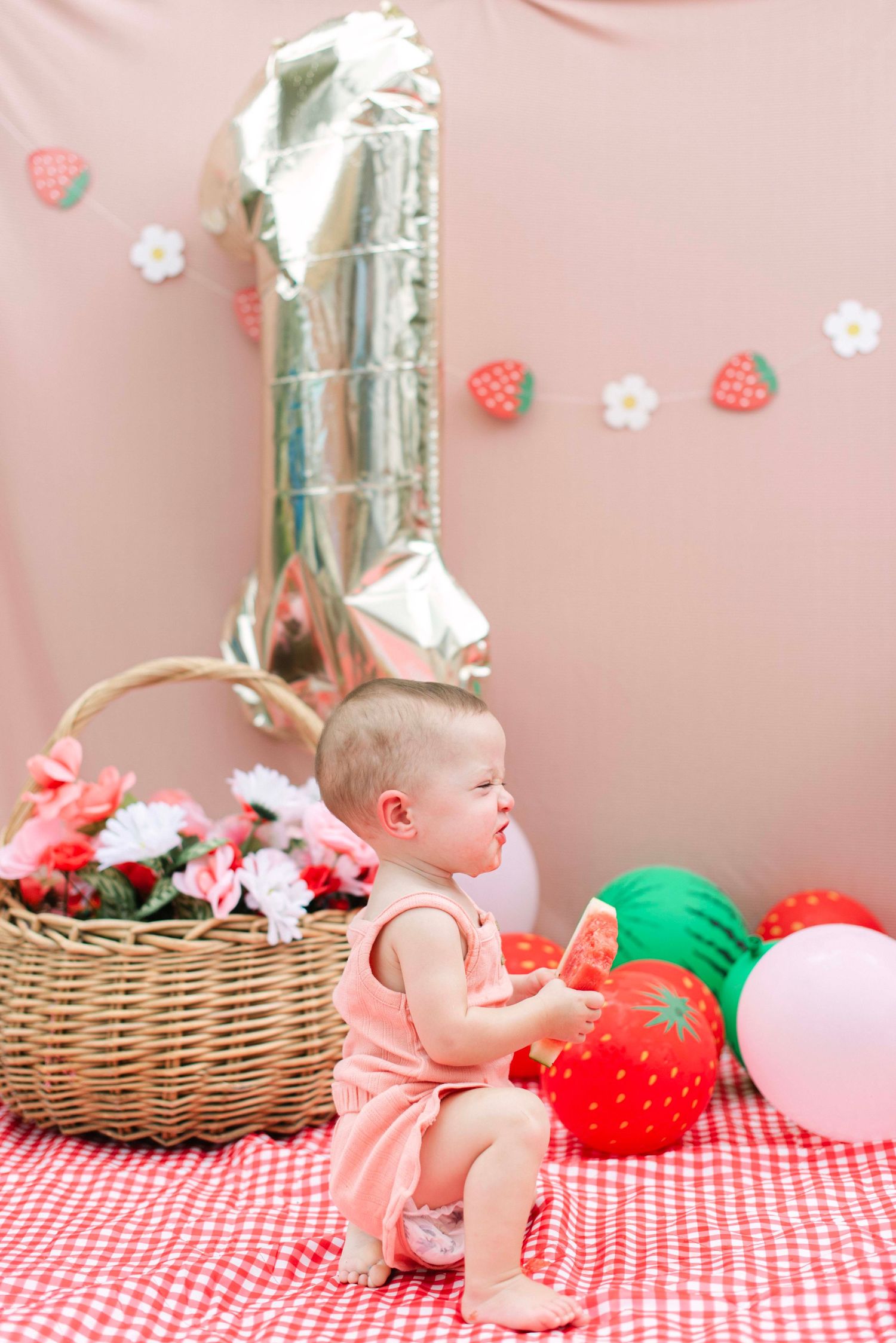 Baby in pink outfit with strawberry themed decorations and balloons on checkered blanket.