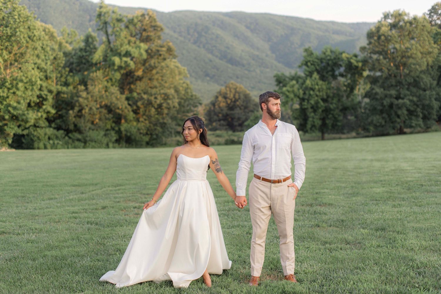 A couple stands together on a lush green lawn with mountain views and trees in the background.