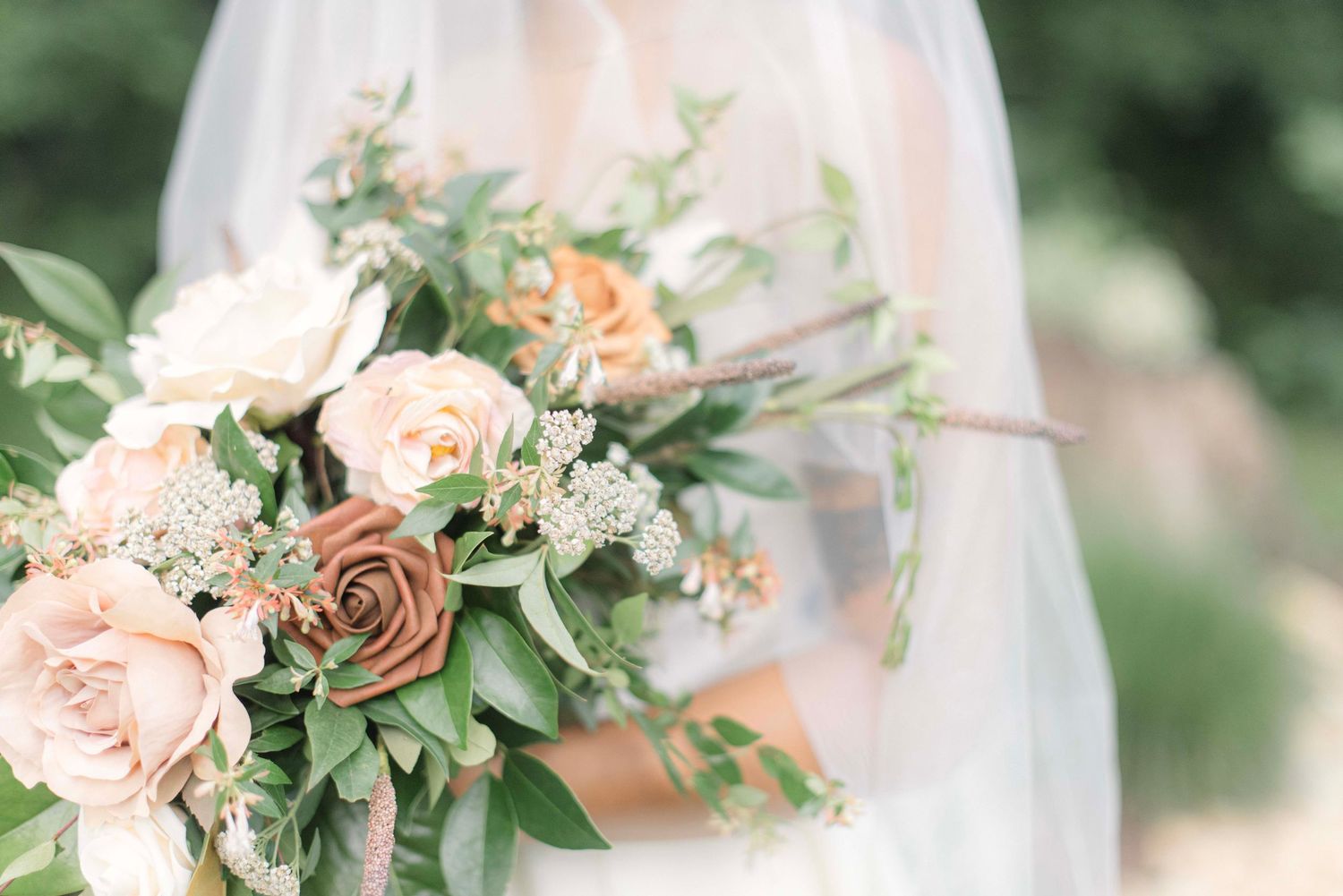 A soft peach and blush wedding bouquet with roses and greenery held against a white dress in natural light.