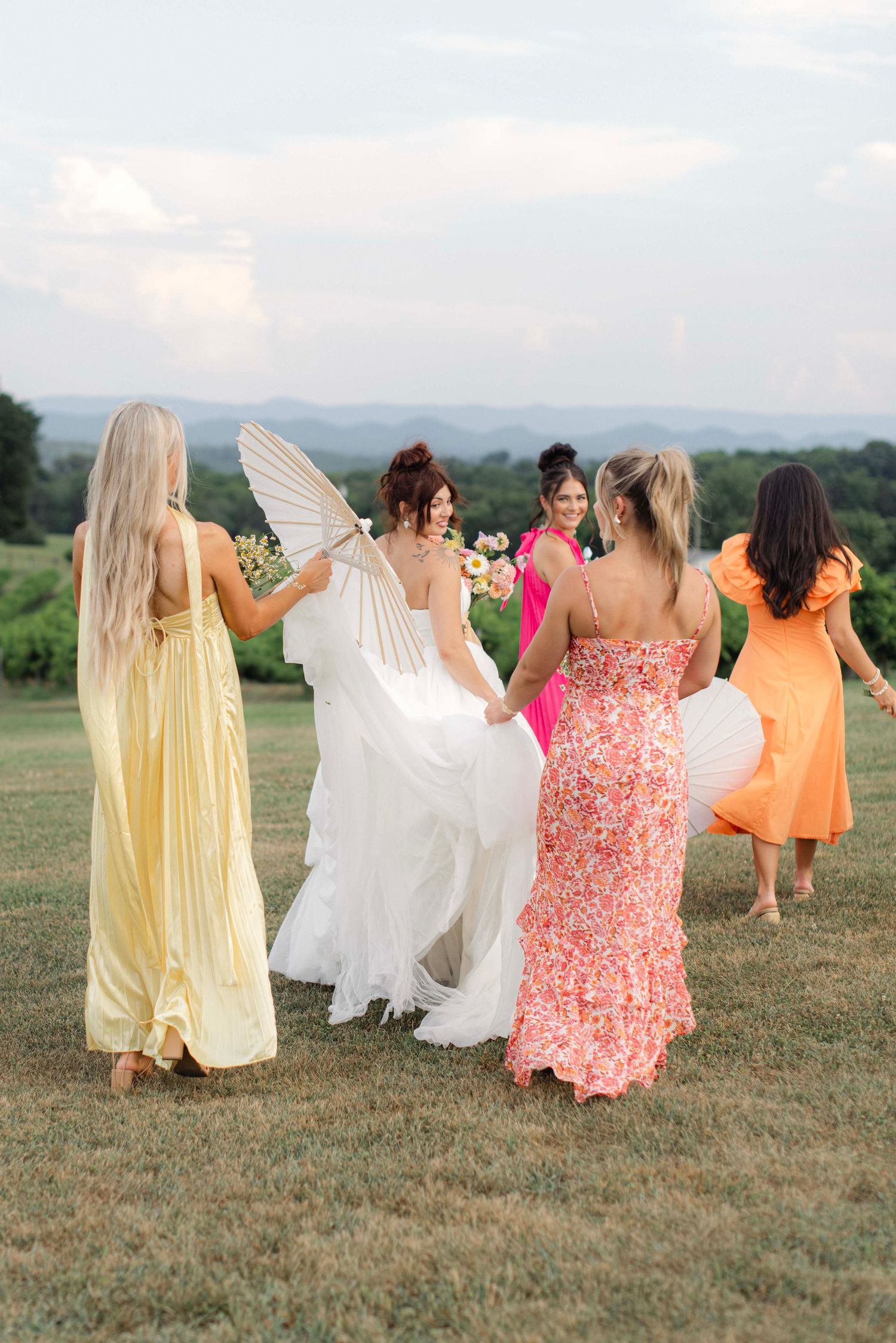 Bridesmaids in colorful dresses dancing and twirling together in a grassy field during sunset.