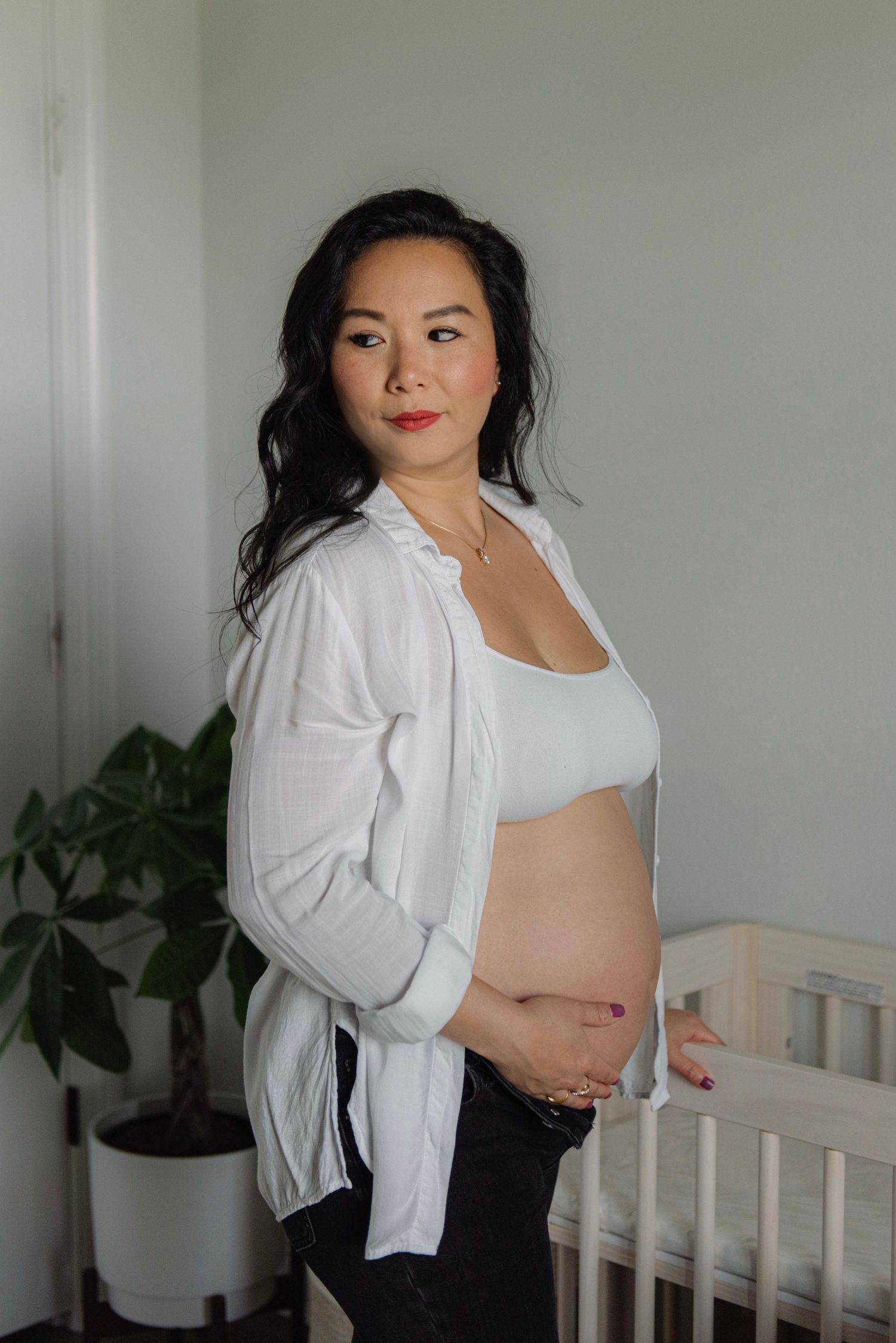 Expectant mother in white cardigan and crop top stands in nursery room with potted plant.