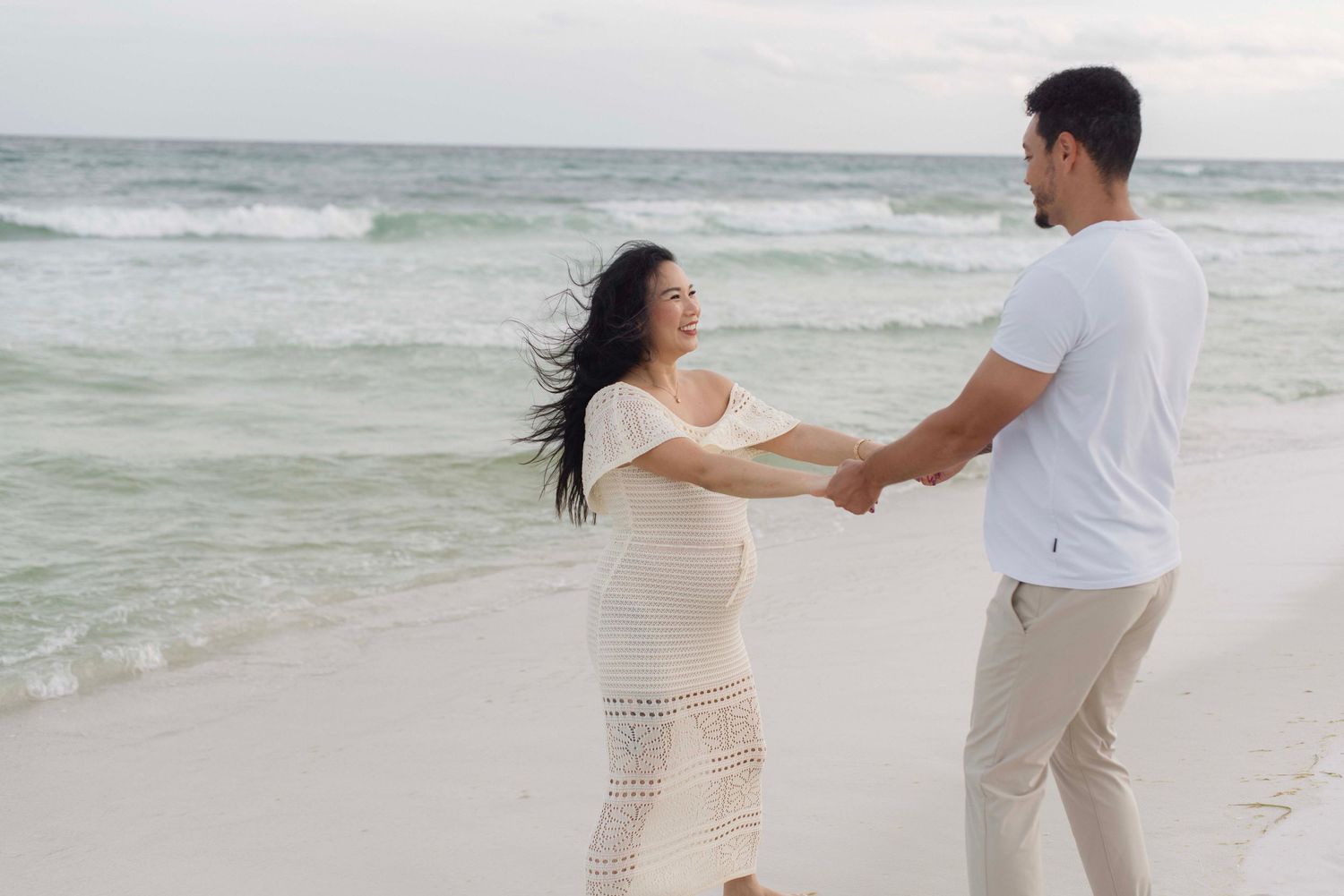 A romantic couple embracing on a sandy beach with ocean waves and cloudy skies behind them.