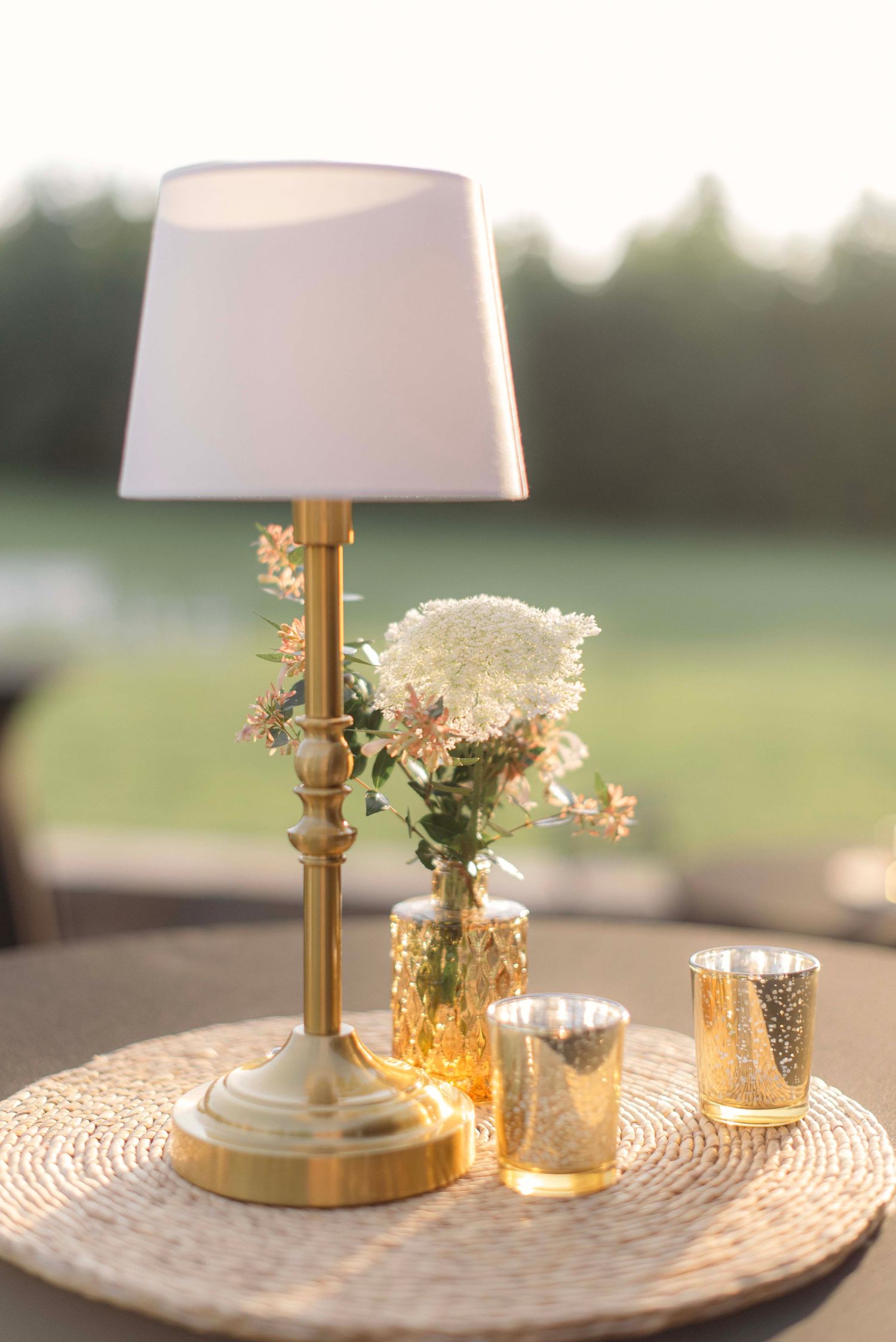 A decorative gold table lamp with white shade sits on a textured round placemat with glowing candle votives at dusk.