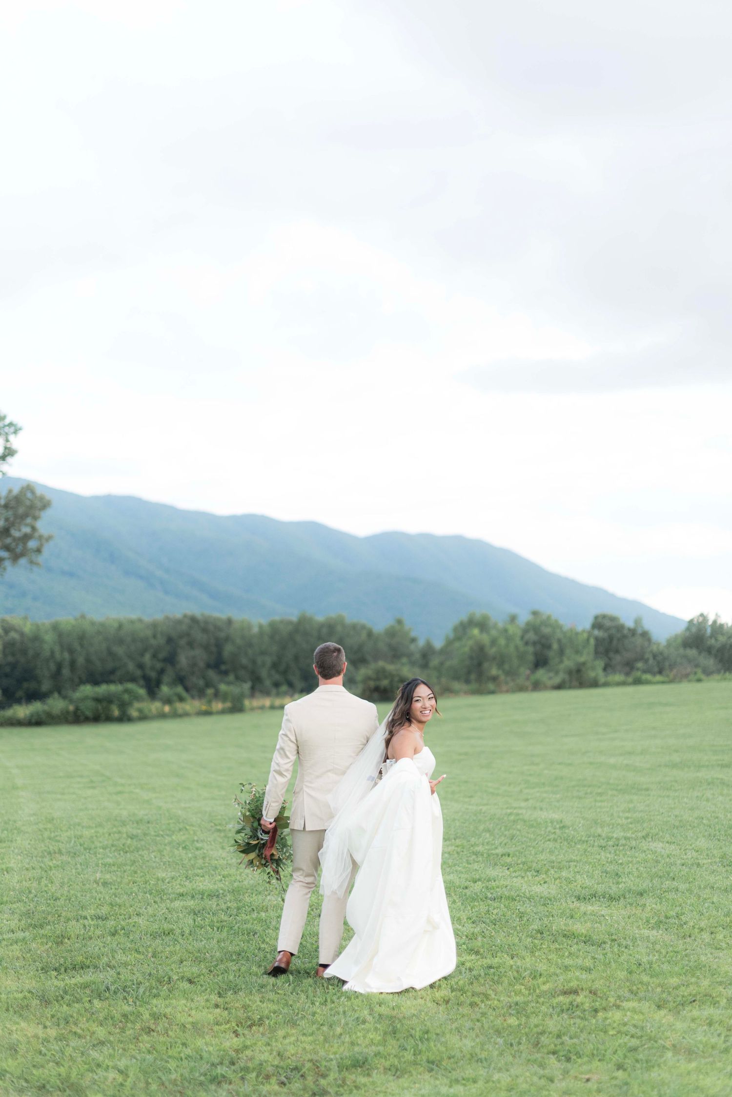 Wedding couple embraces in scenic mountain meadow with rolling green hills and distant peaks as backdrop.