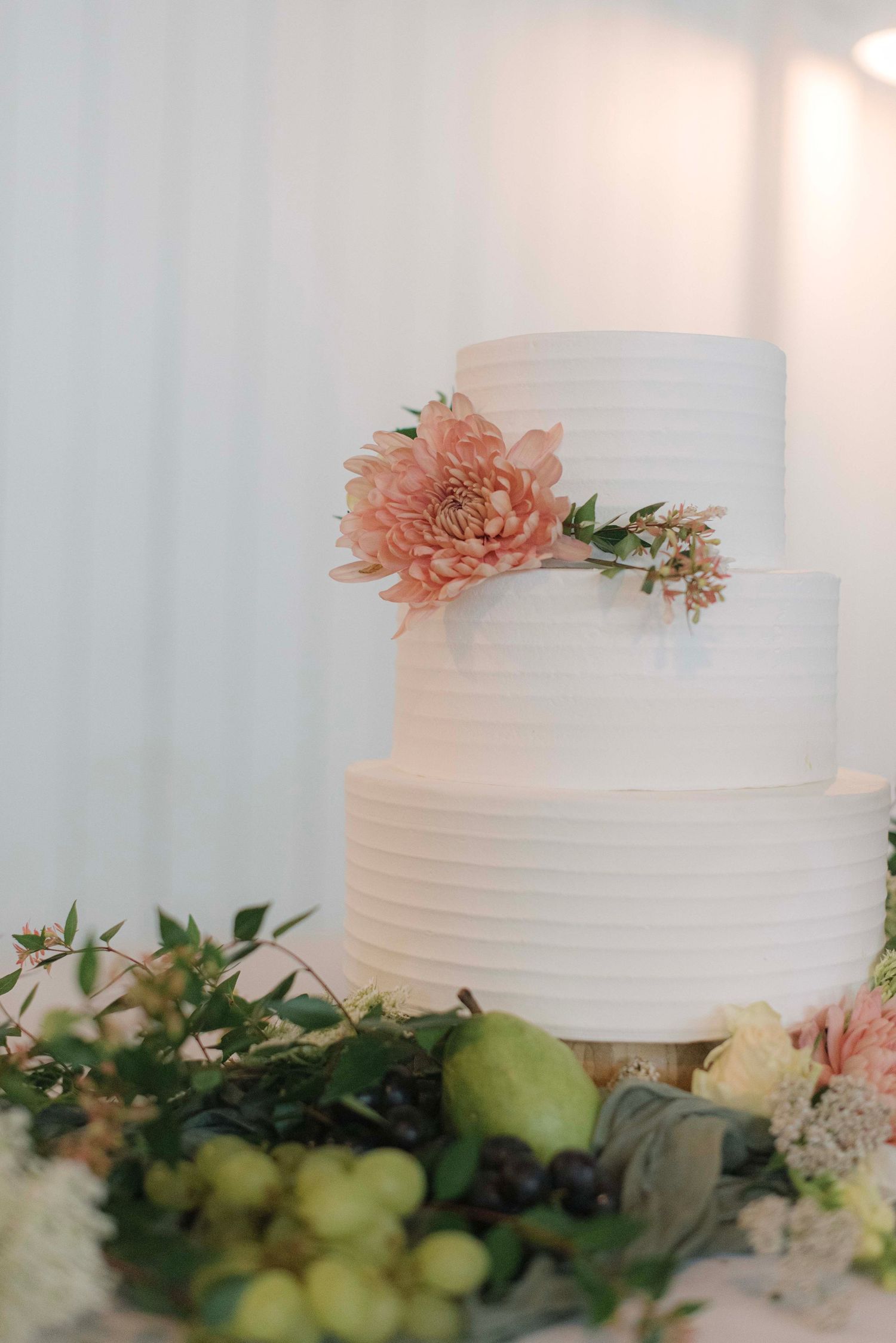 Elegant white tiered wedding cake decorated with a peach-colored flower and greenery, displayed with fresh fruit and foliage.