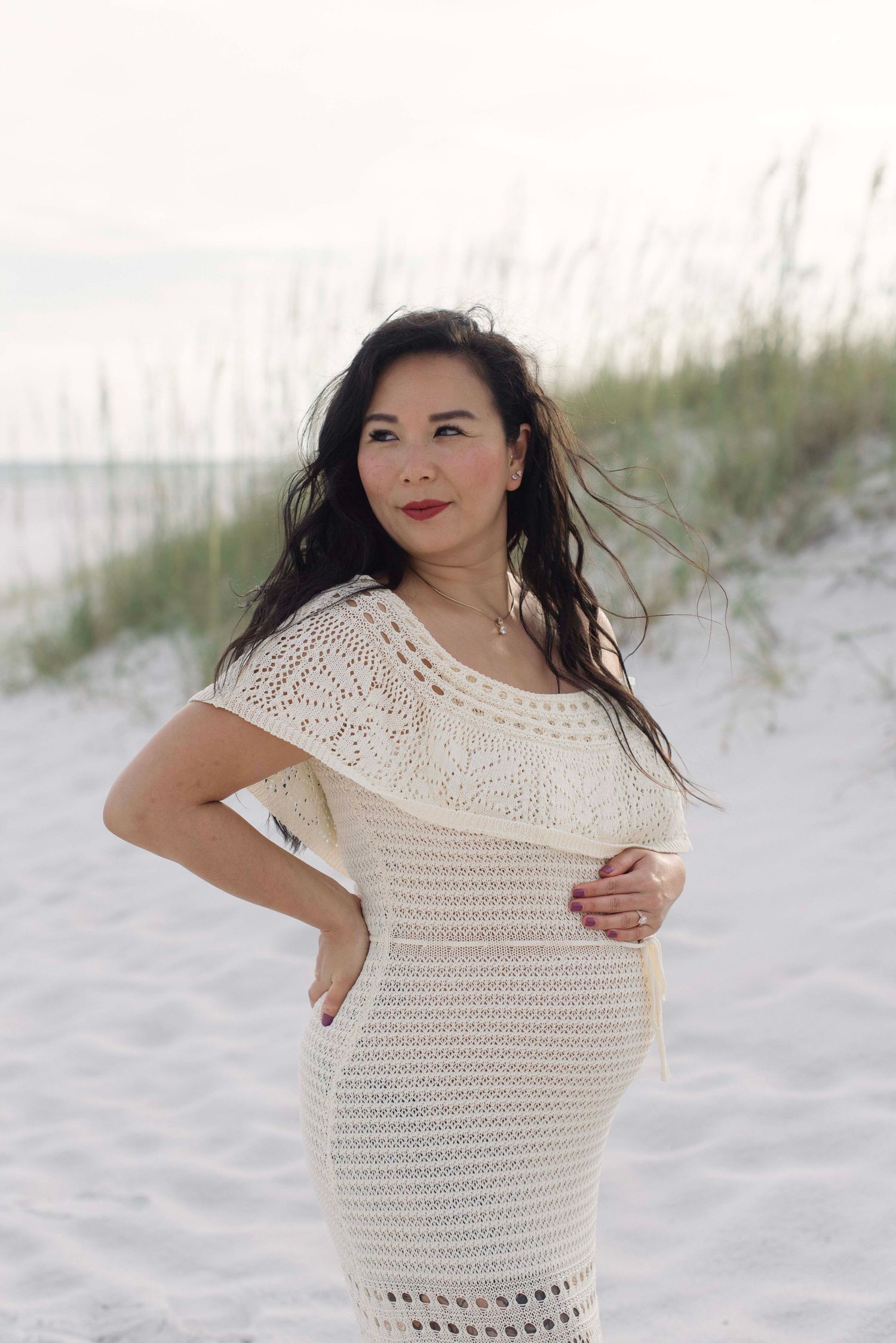 Candid beach maternity portraits showing a woman in a delicate white crocheted dress against sand dunes.
