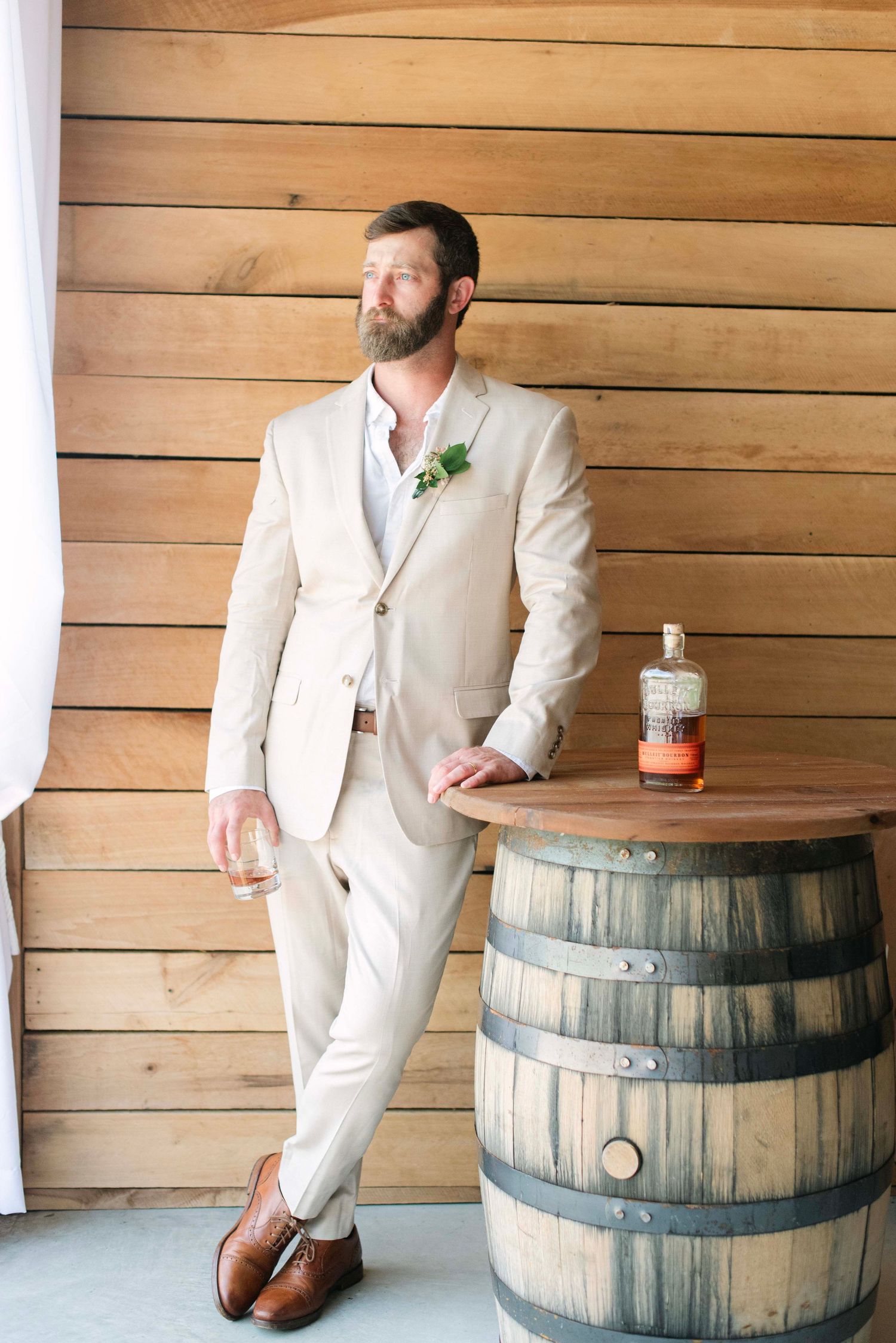 A person in a white suit leans casually against wooden barrels in a rustic setting with wood-paneled walls.