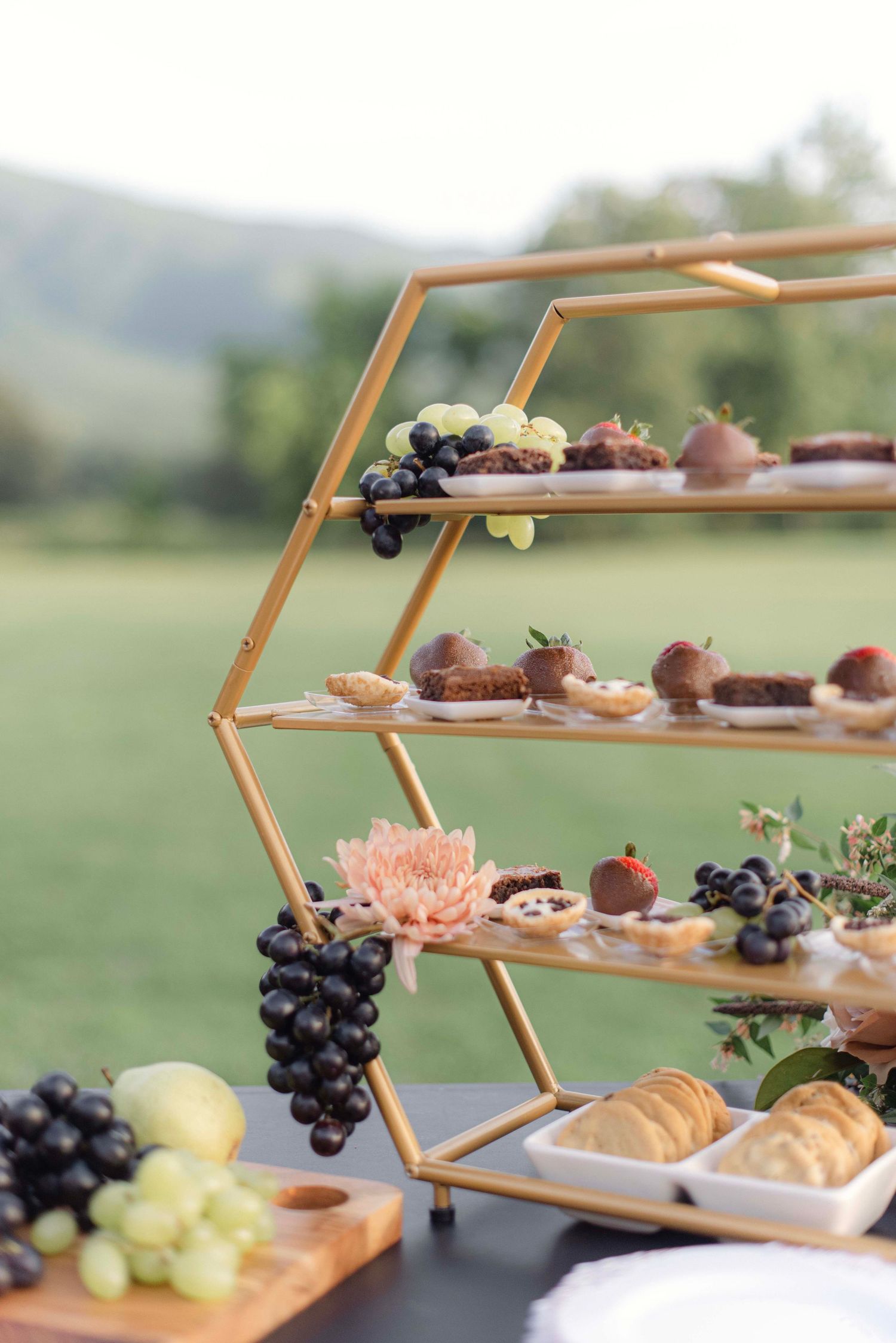 Wooden hexagonal tiered dessert stand displaying pastries, grapes, and fruit at an outdoor event.