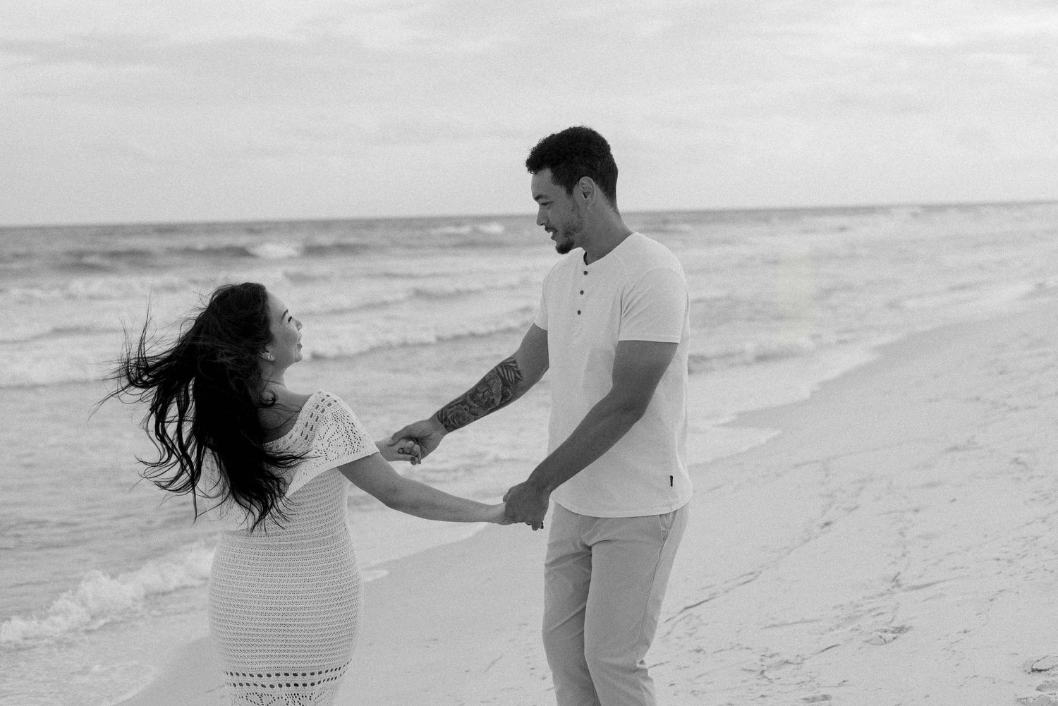 A couple walks and plays together on a sandy beach at sunset in black and white.