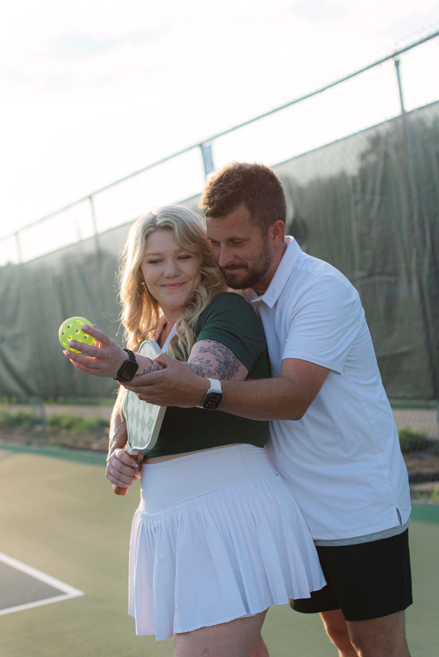 A couple shares an intimate moment on a tennis court at sunset while holding tennis balls.