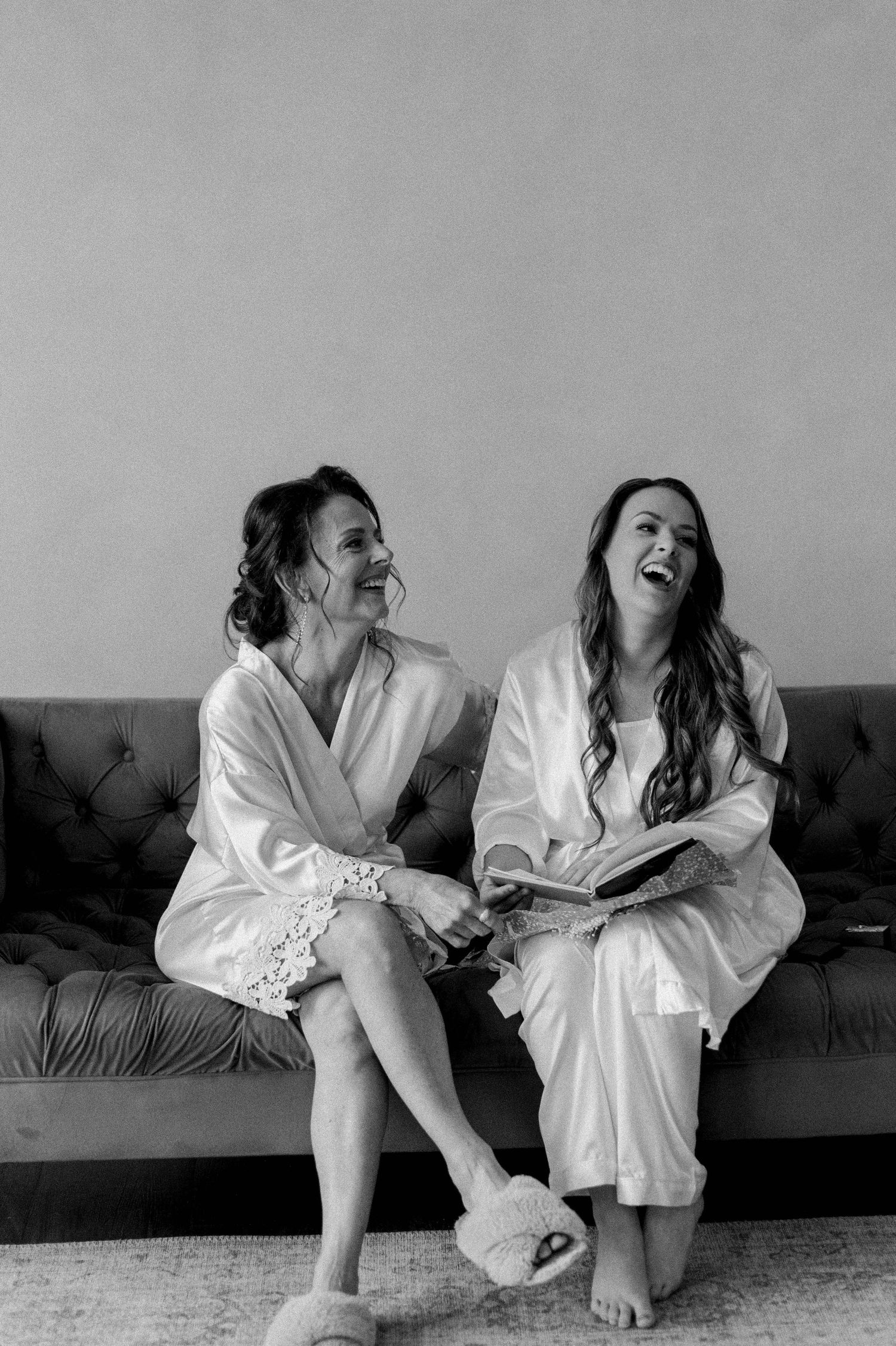 Women in white robes lounge casually on a dark couch during a relaxed photoshoot.