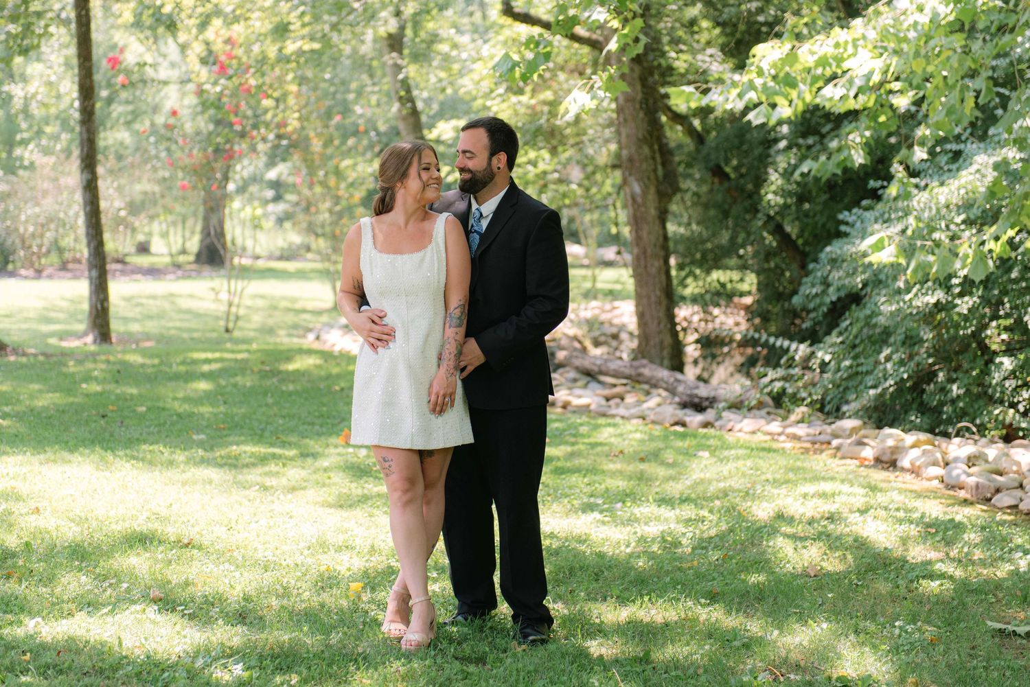Couple in formal attire walking through a lush green garden setting on a sunny day.