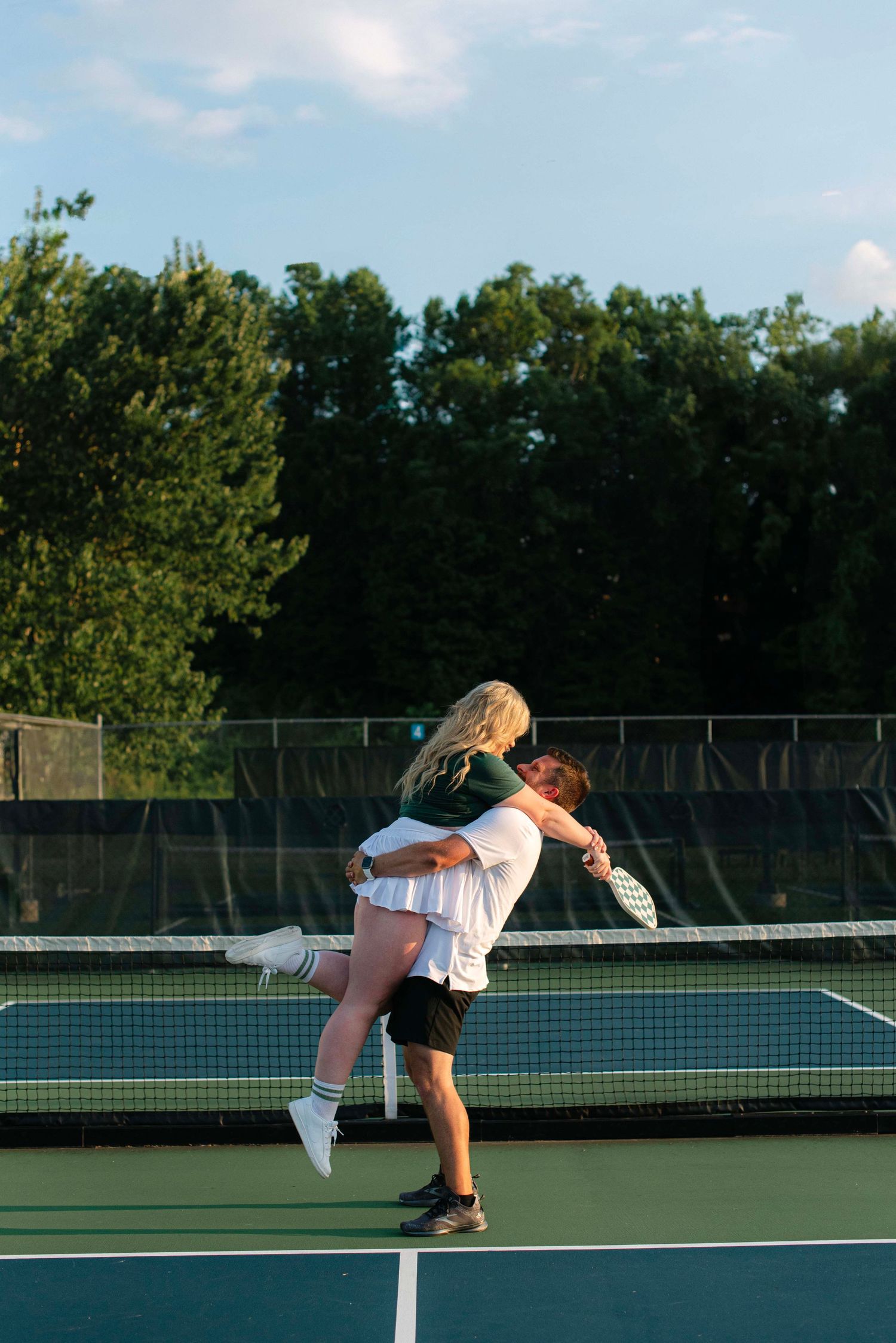 A couple embraces playfully on an outdoor tennis court during sunset with trees in the background.
