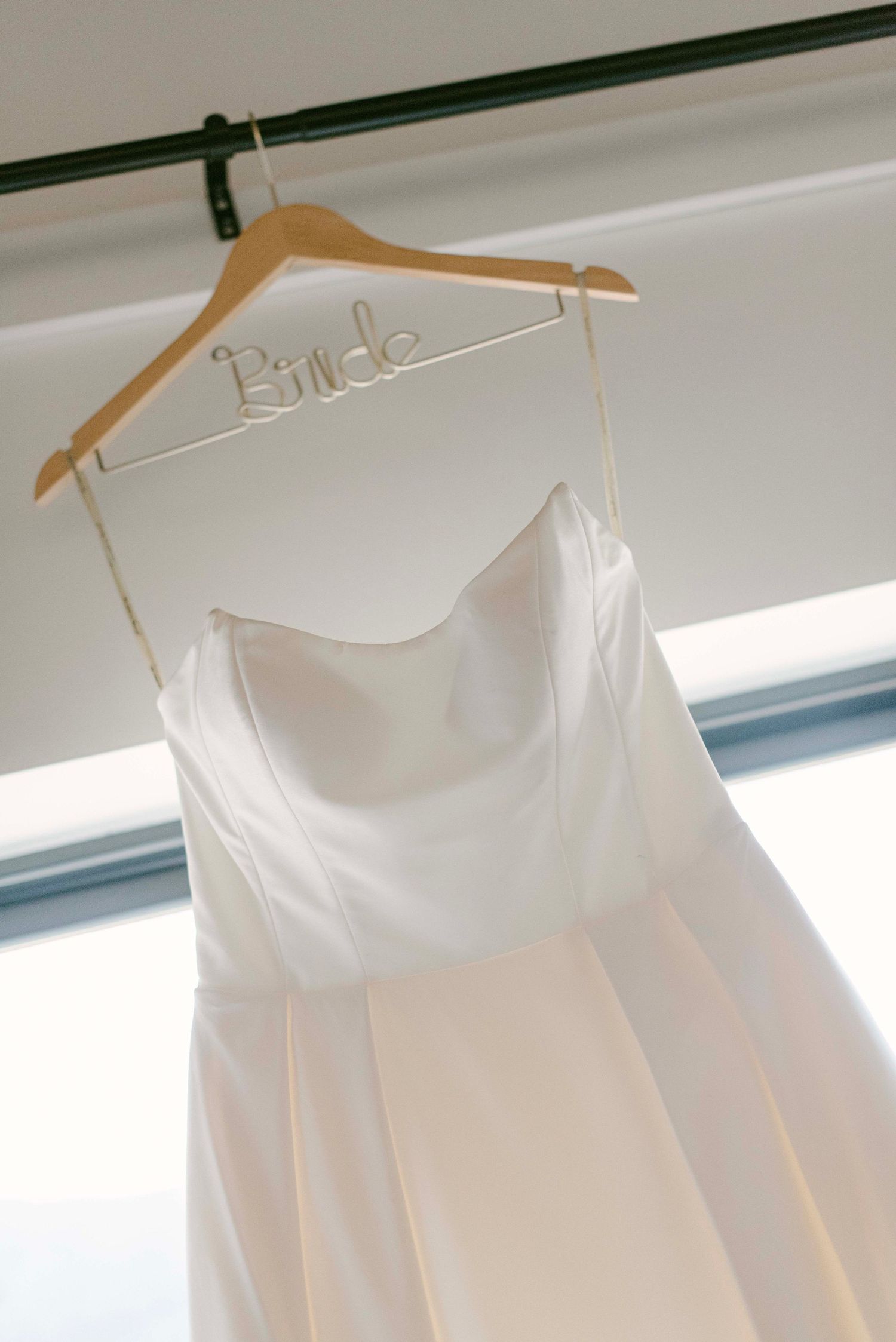 A white wedding dress hangs on a wooden hanger labeled 'Bride' against a light background.