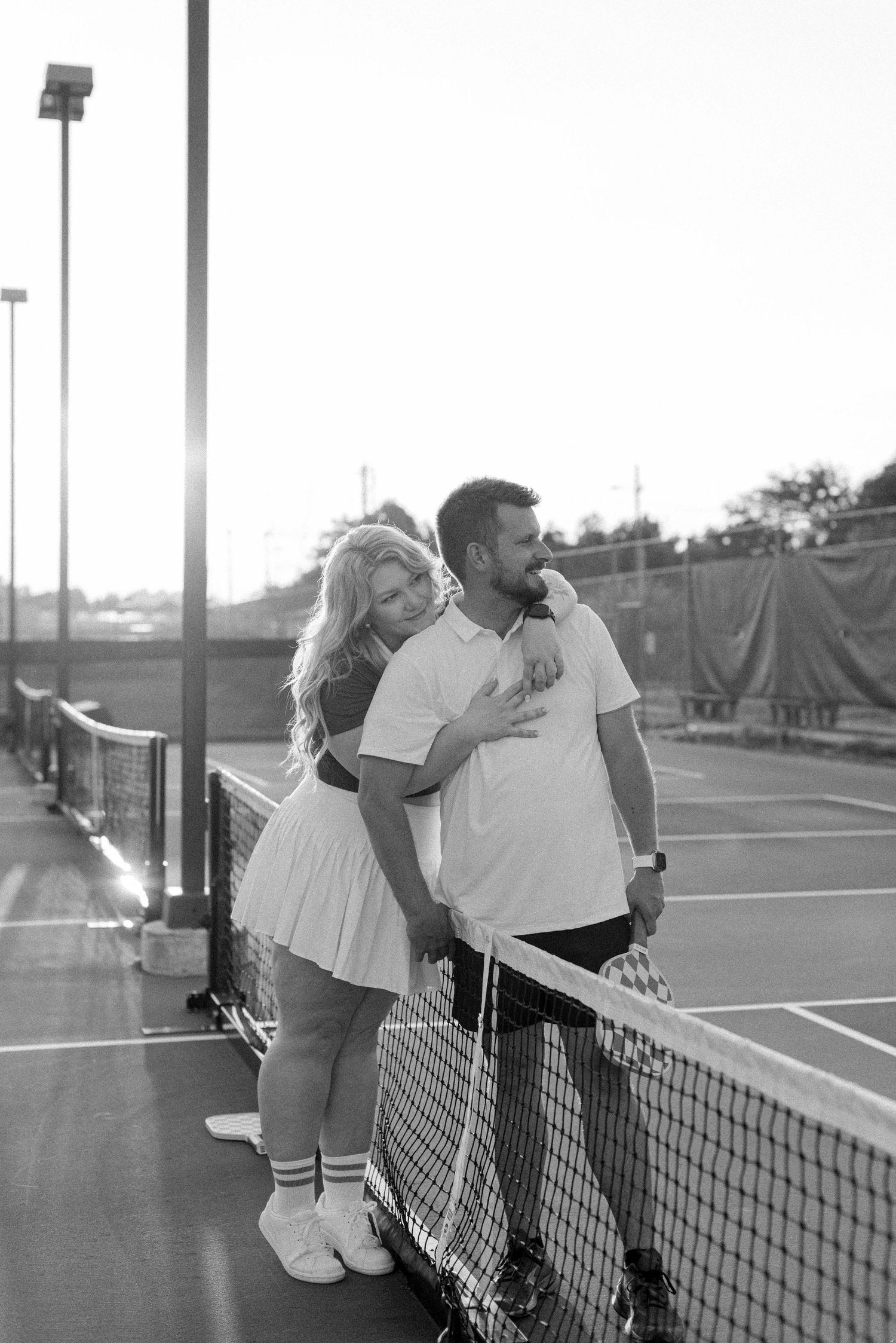 A couple shares an intimate moment on a tennis court in black and white, leaning against the net.