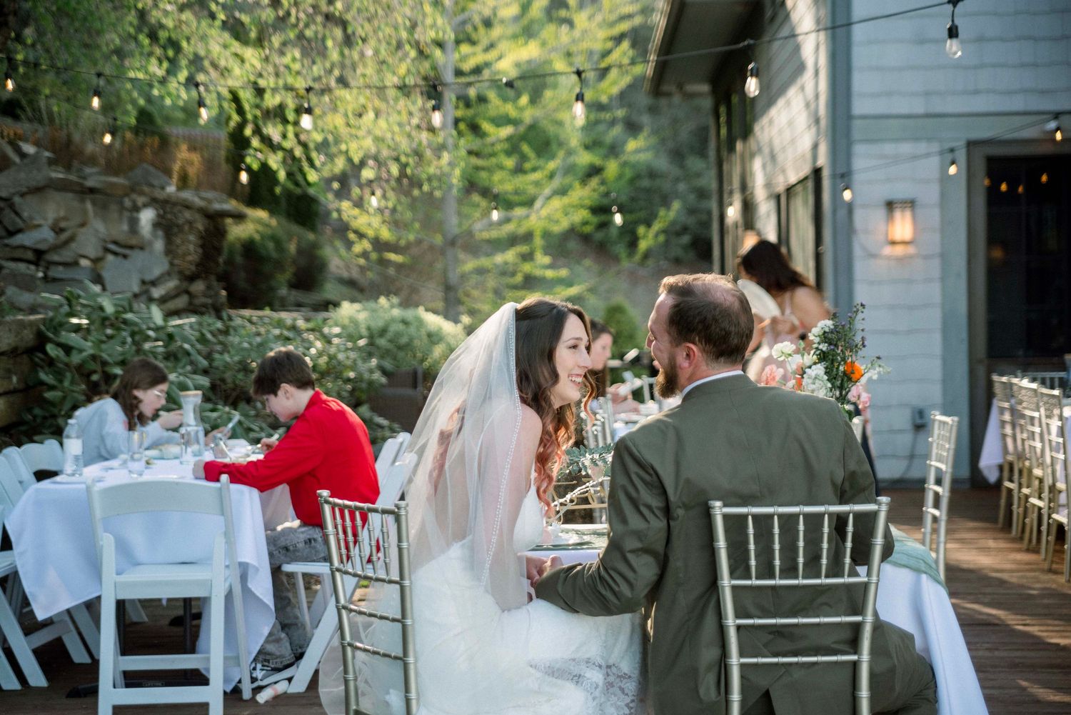 Outdoor wedding reception setup with white tables and chairs under trees.