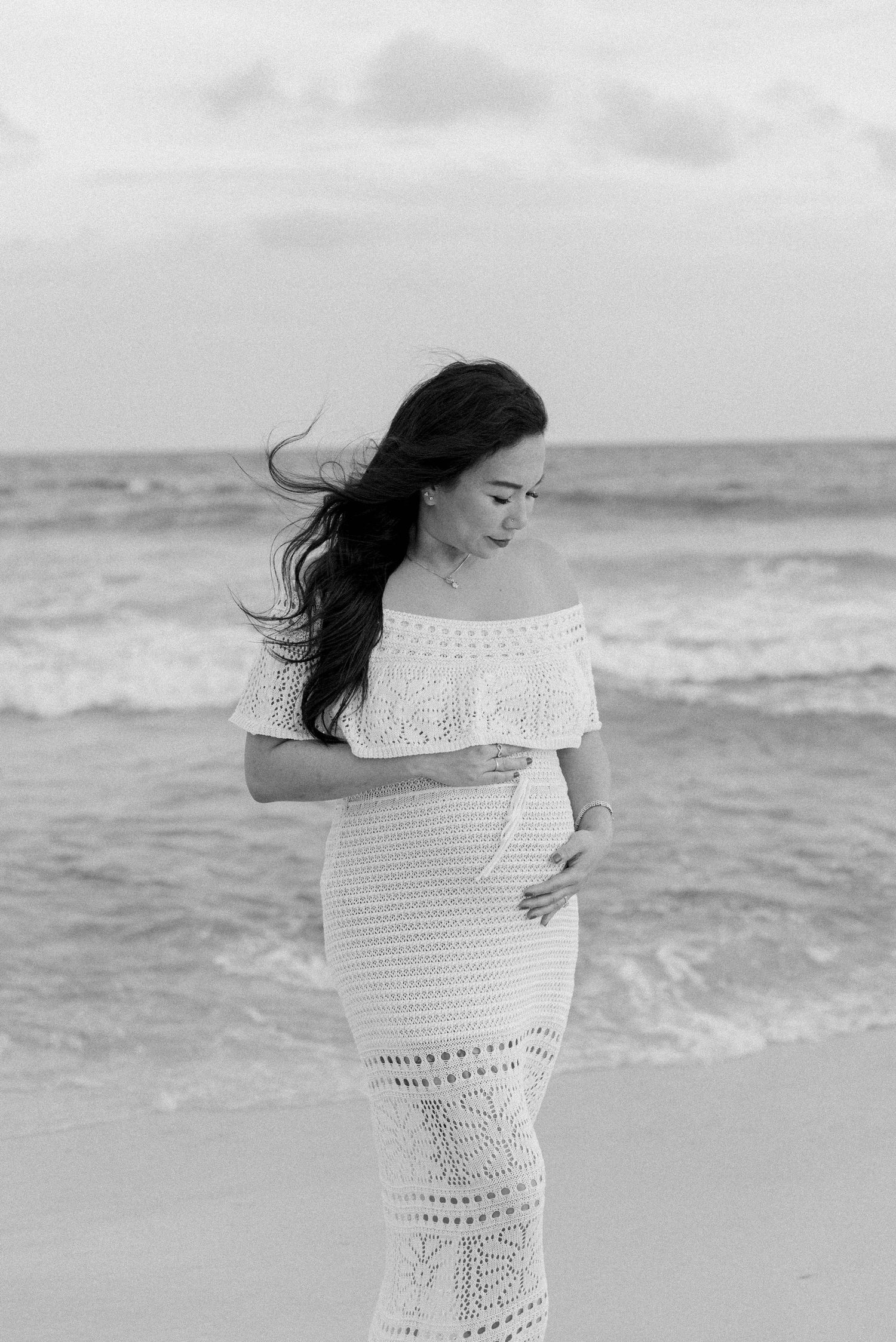 A person in a white crochet dress stands on a beach while their dark hair blows in the wind.