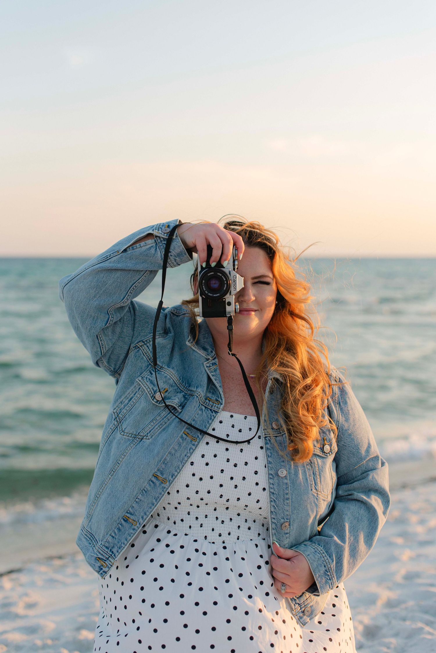 Photographer in polka dot dress and denim jacket shooting at beach during sunset.