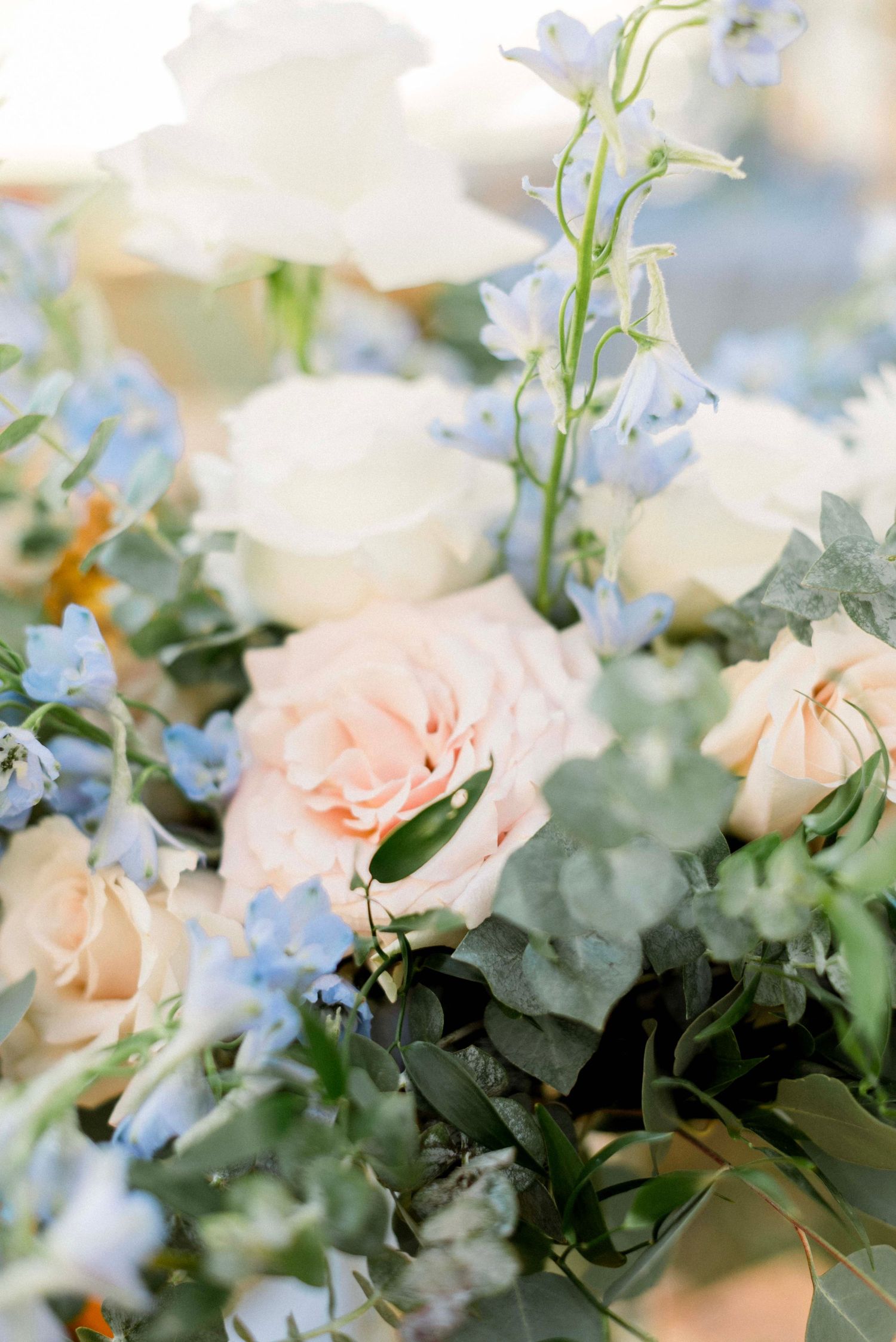 Delicate floral arrangement with pale pink roses and blue flowers mixed with green foliage in soft focus.