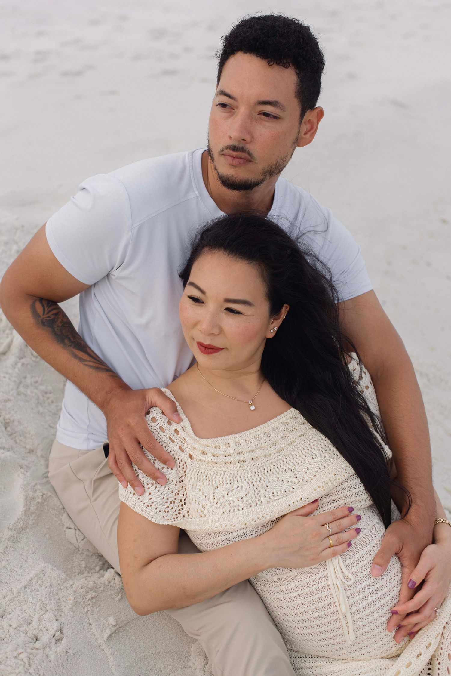 A romantic couple embraces on a sandy beach, with the woman wearing a white crochet dress.