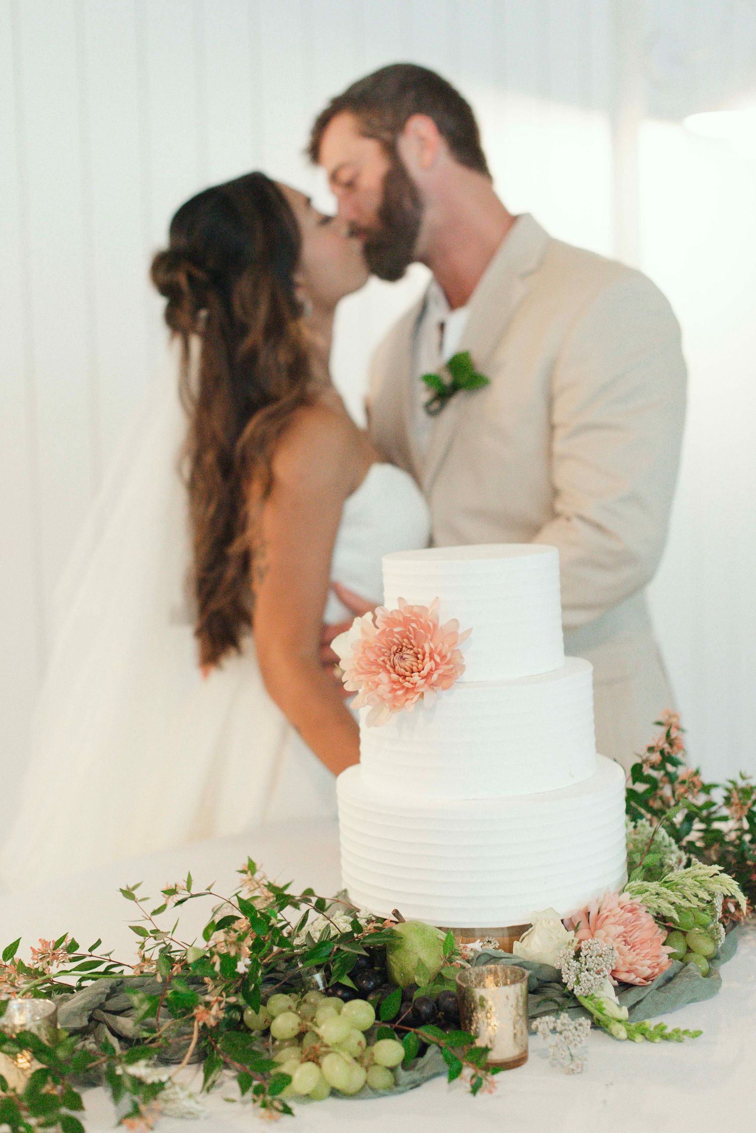 Elegant white wedding cake decorated with peach flowers and greenery sits before a romantic moment at a wedding reception.