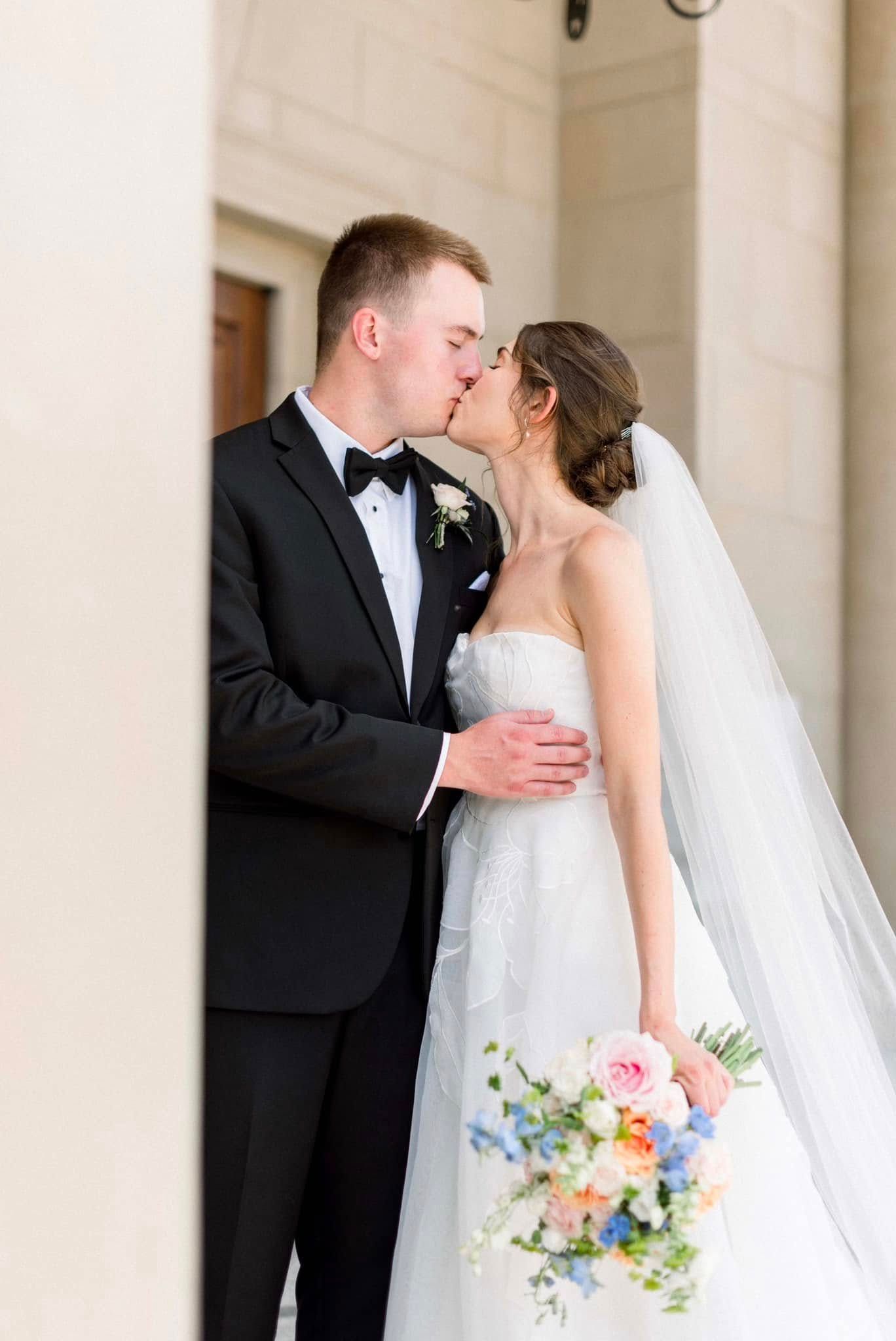 Elegant bride and groom share a romantic kiss under white columns while holding a colorful spring bouquet.