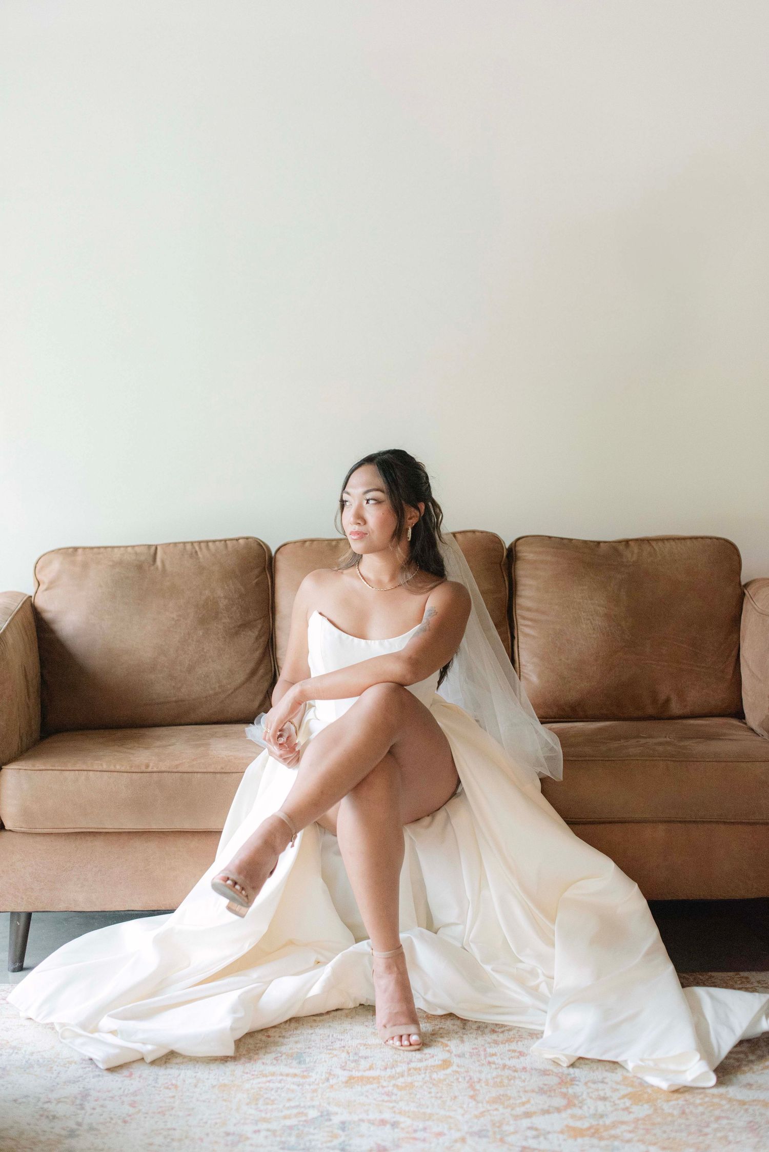 A bride in a white wedding dress reclines on a brown leather couch in an artistic boudoir photo series.