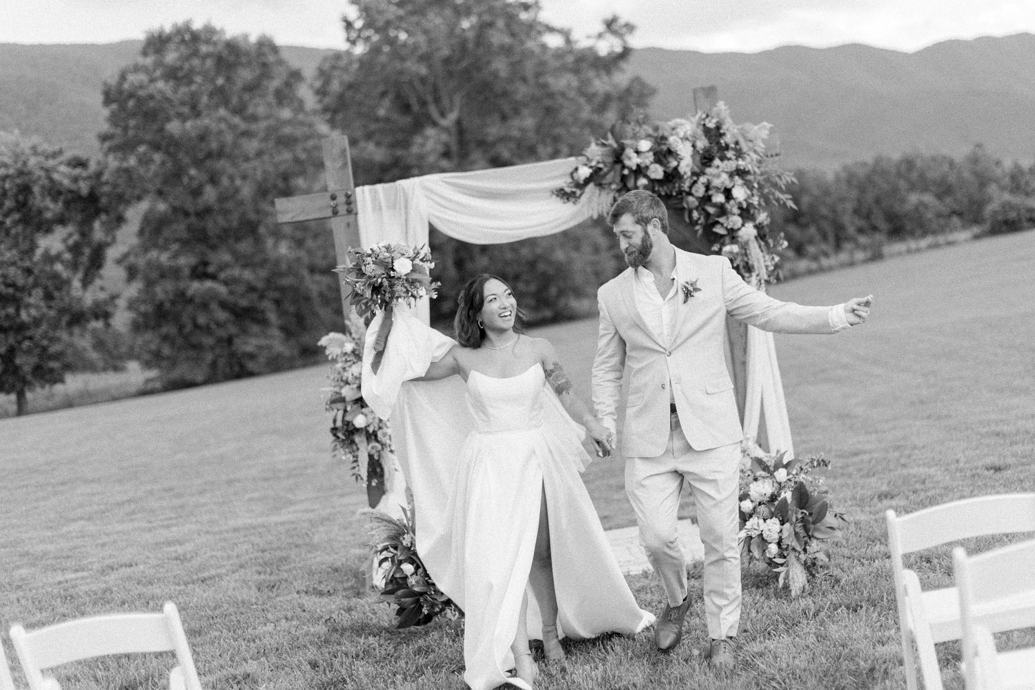 A joyful couple celebrates during their outdoor wedding ceremony beneath a floral arch on a hillside.