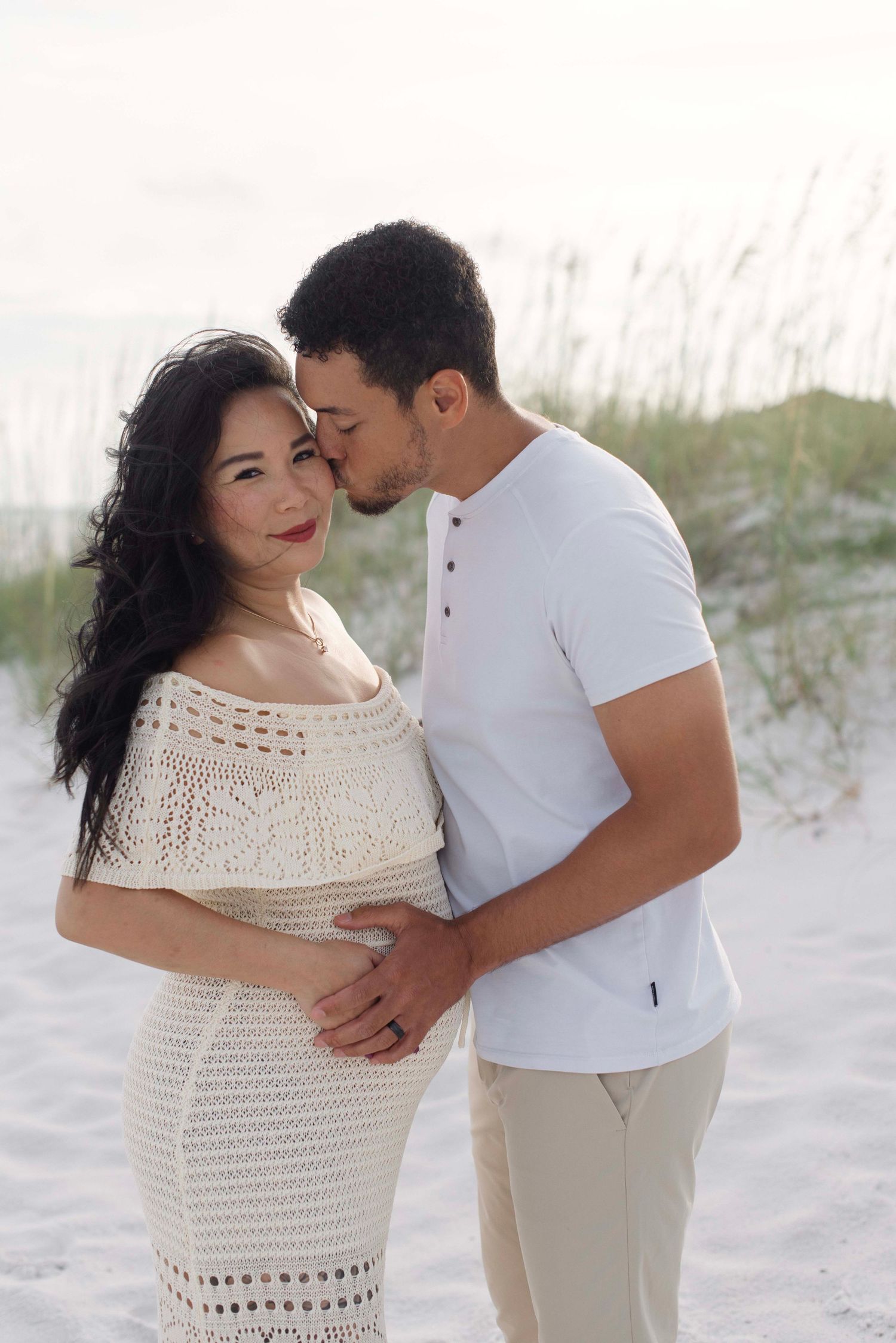 A romantic couple embraces on a white sandy beach with sea grass in the background during a photoshoot.