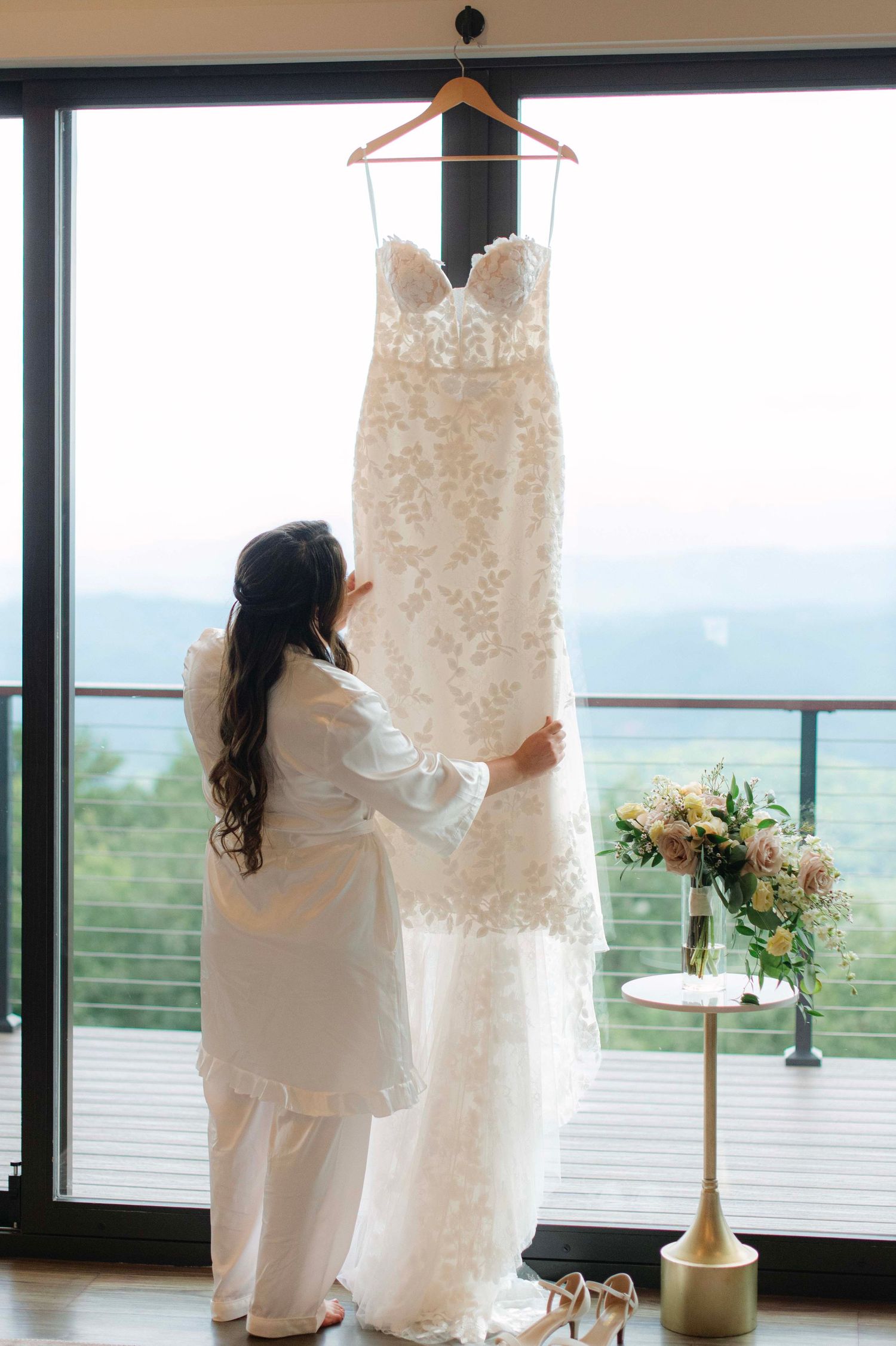 A lace wedding dress hangs by a window with scenic mountain views in the background.