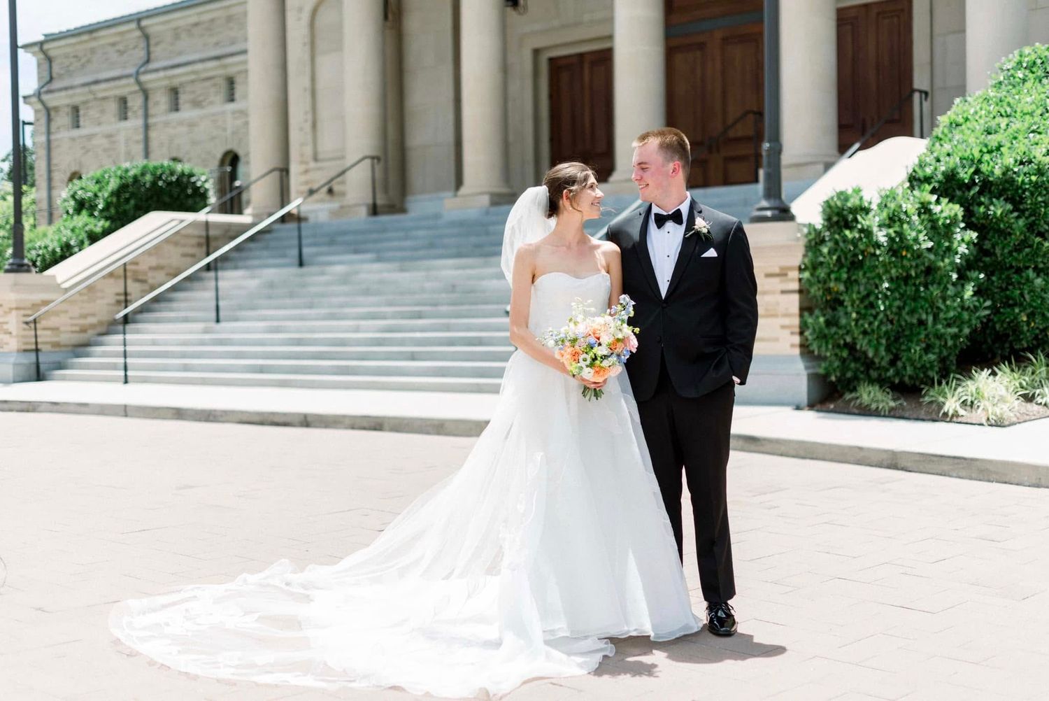 A bride and groom pose for wedding photos on grand stone steps outside an elegant classical building.