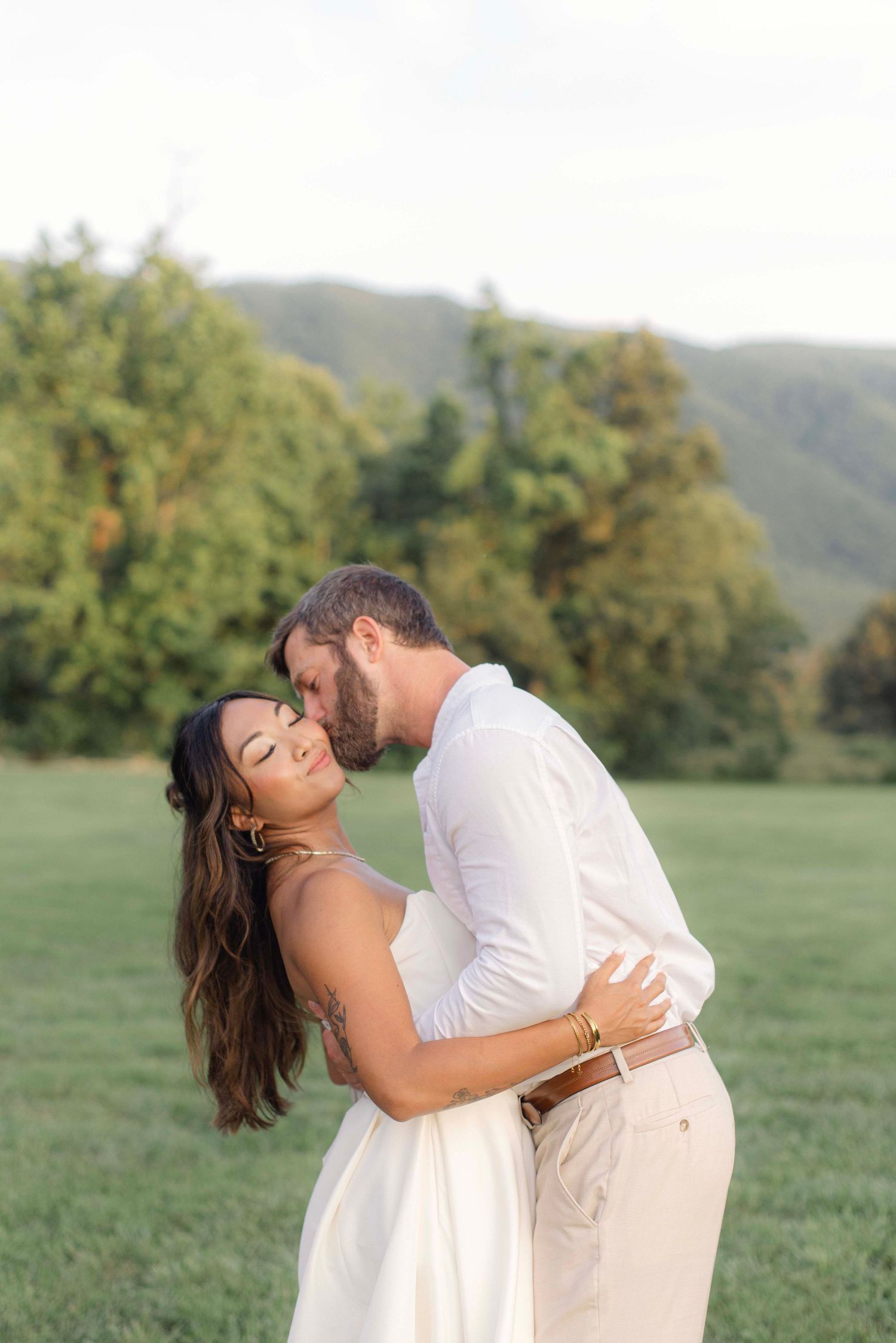 A couple in white attire shares a romantic dip pose in a sunny meadow.