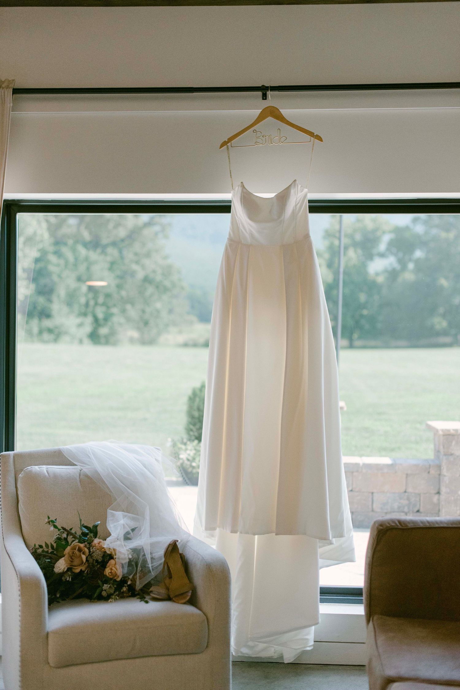 White wedding dresses hanging on hangers against large windows in a bright, sunlit dressing room.