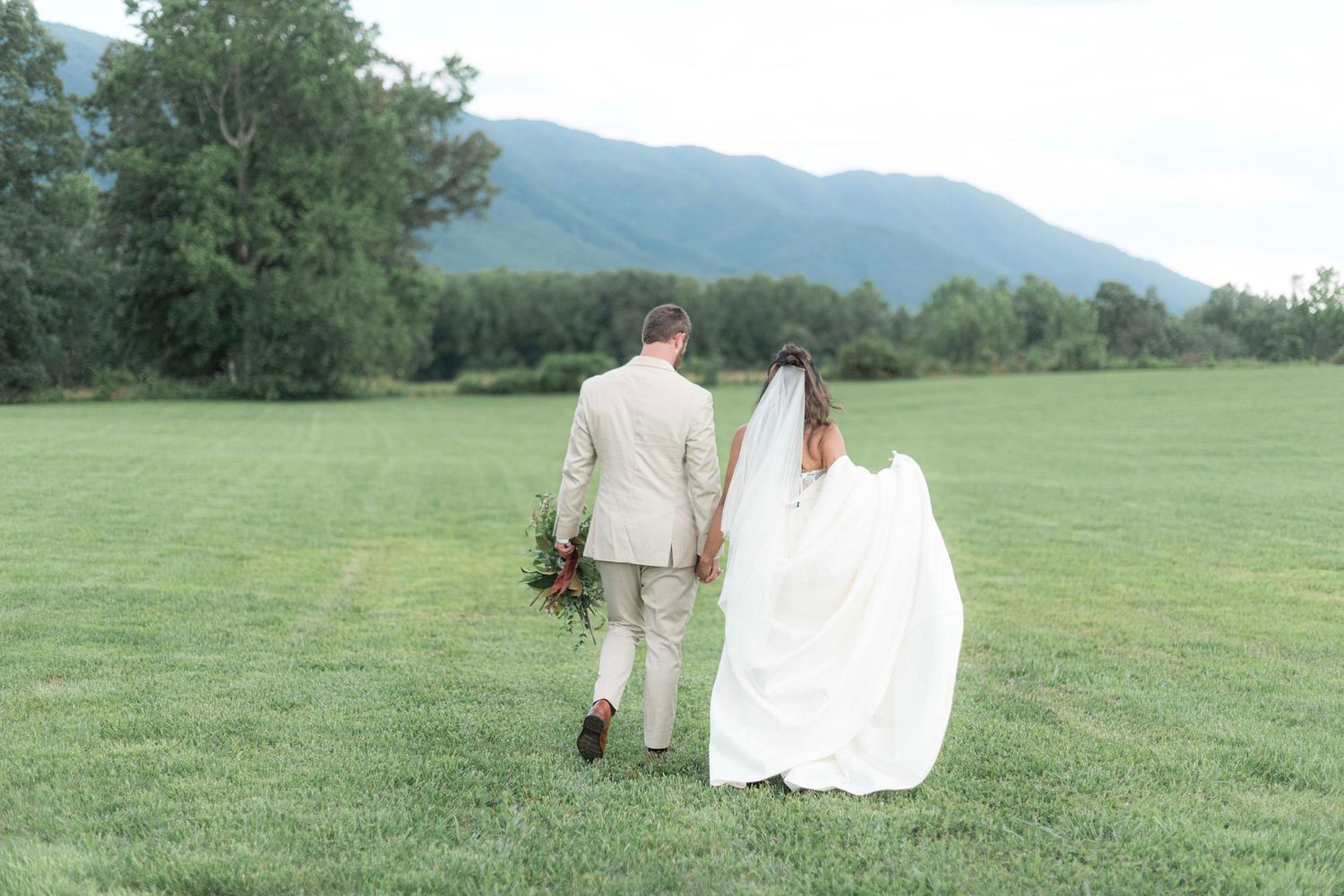 A bride and groom walk hand in hand across a lush green field with mountains in the background.