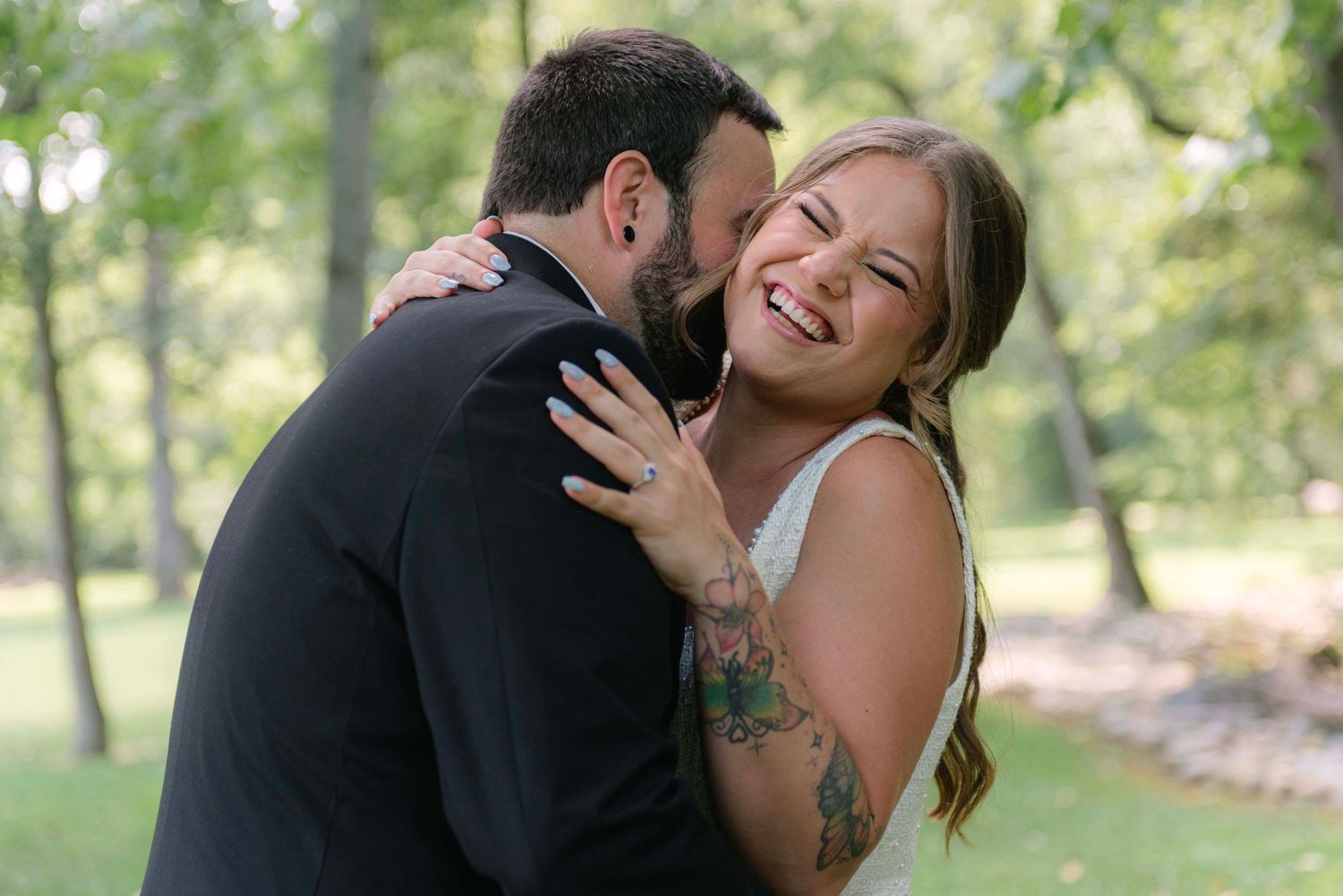 A joyful couple embraces and laughs together while taking photos in a sunlit wooded area.