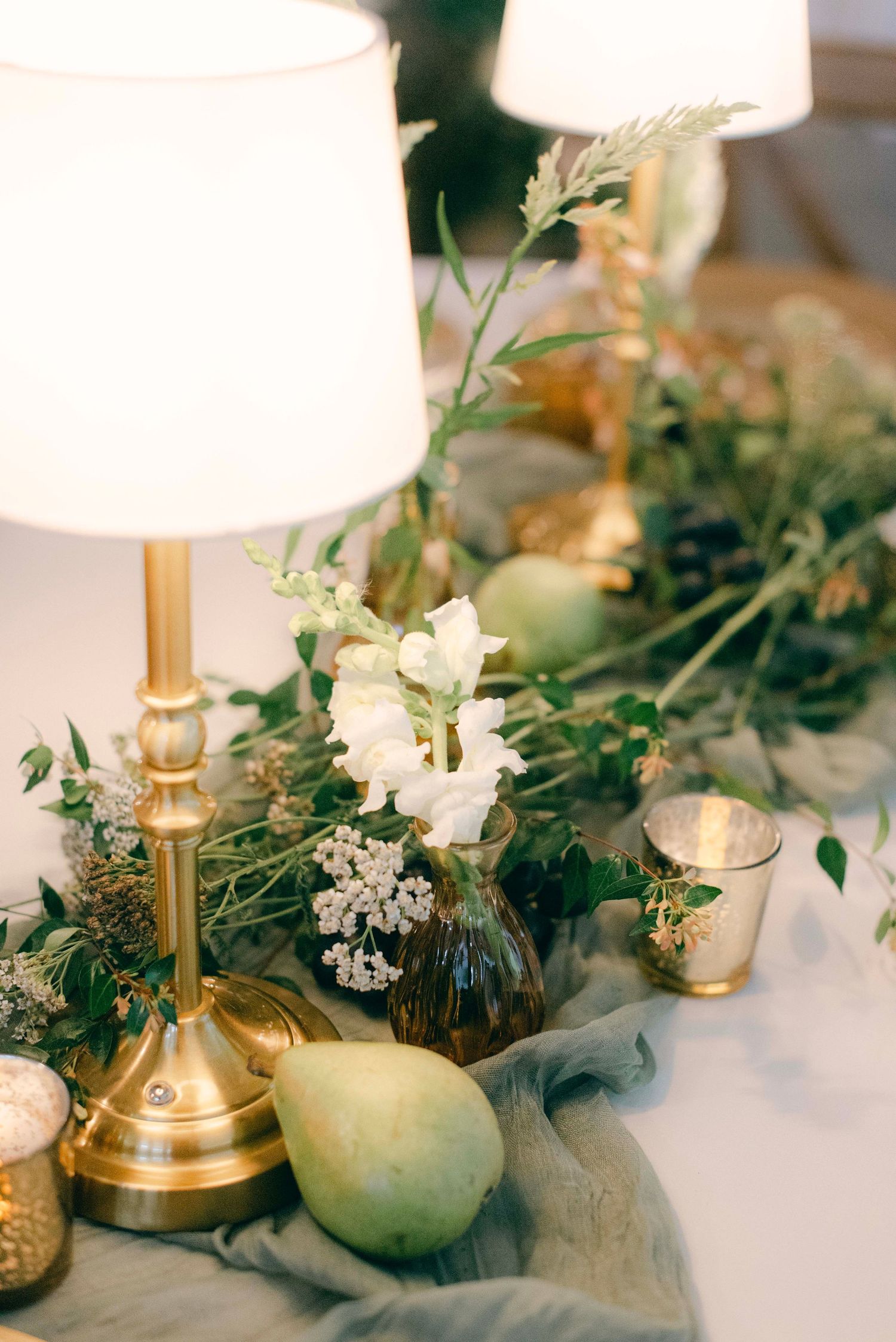 Elegant table decor with brass lamp, white flowers, pear, and greenery arrangements on soft gray linen.