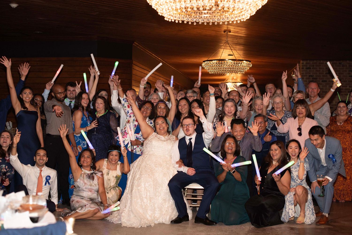 Large group of wedding guests cheering and celebrating on a dance floor under chandeliers.