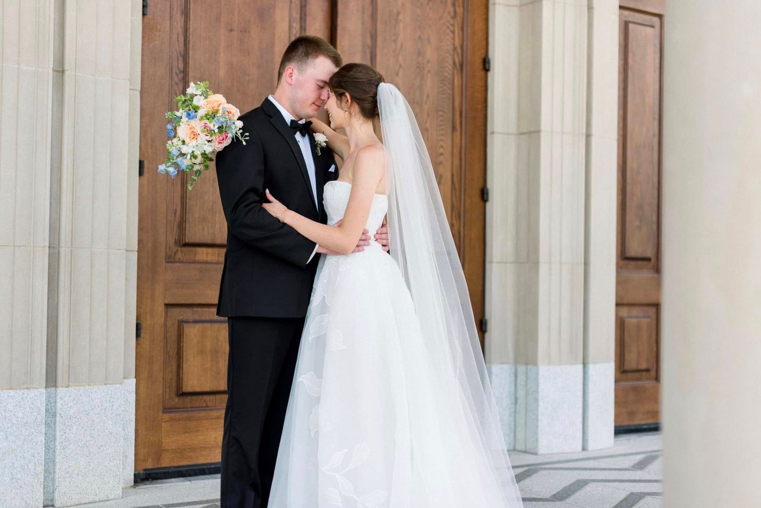 A bride and groom share an intimate moment during their wedding ceremony in front of elegant wooden church doors.