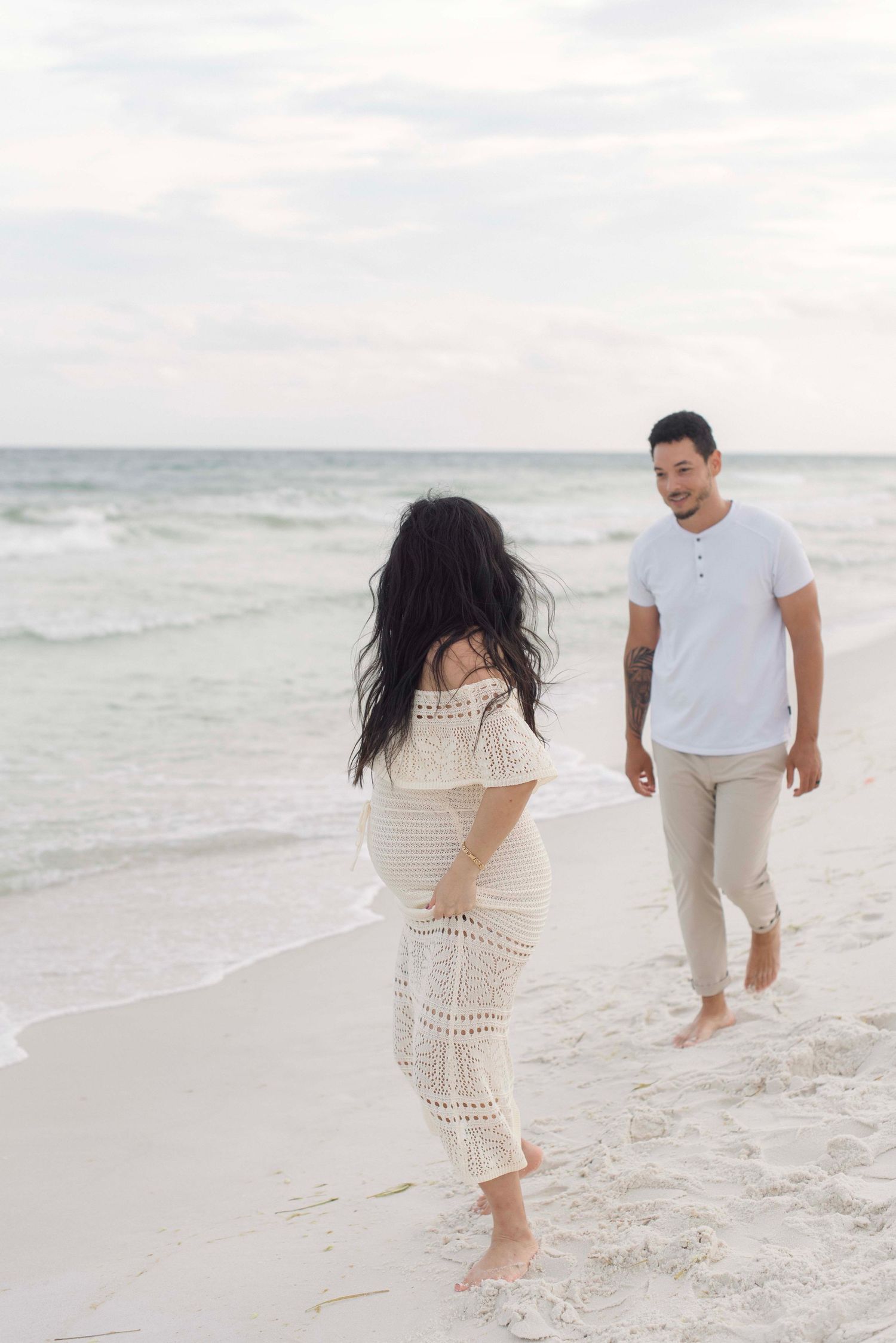 A couple walking barefoot on a white sandy beach at sunset wearing light colored casual beach attire.