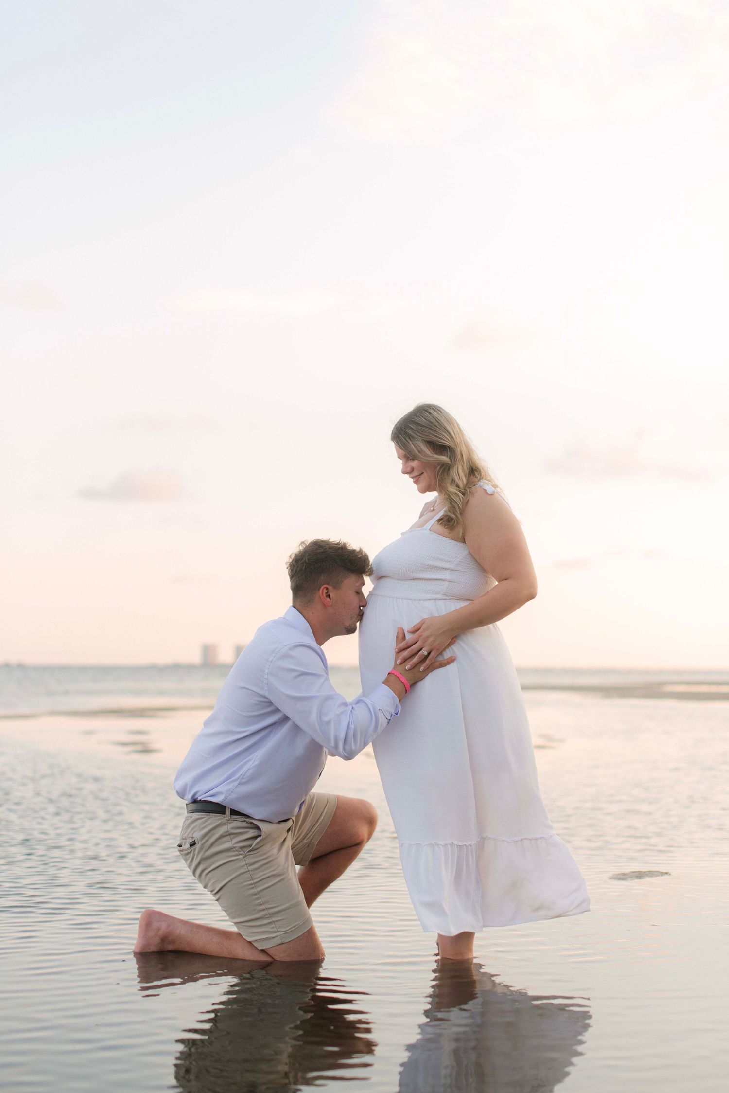 A romantic couple poses in shallow ocean water at sunset wearing white and khaki outfits.