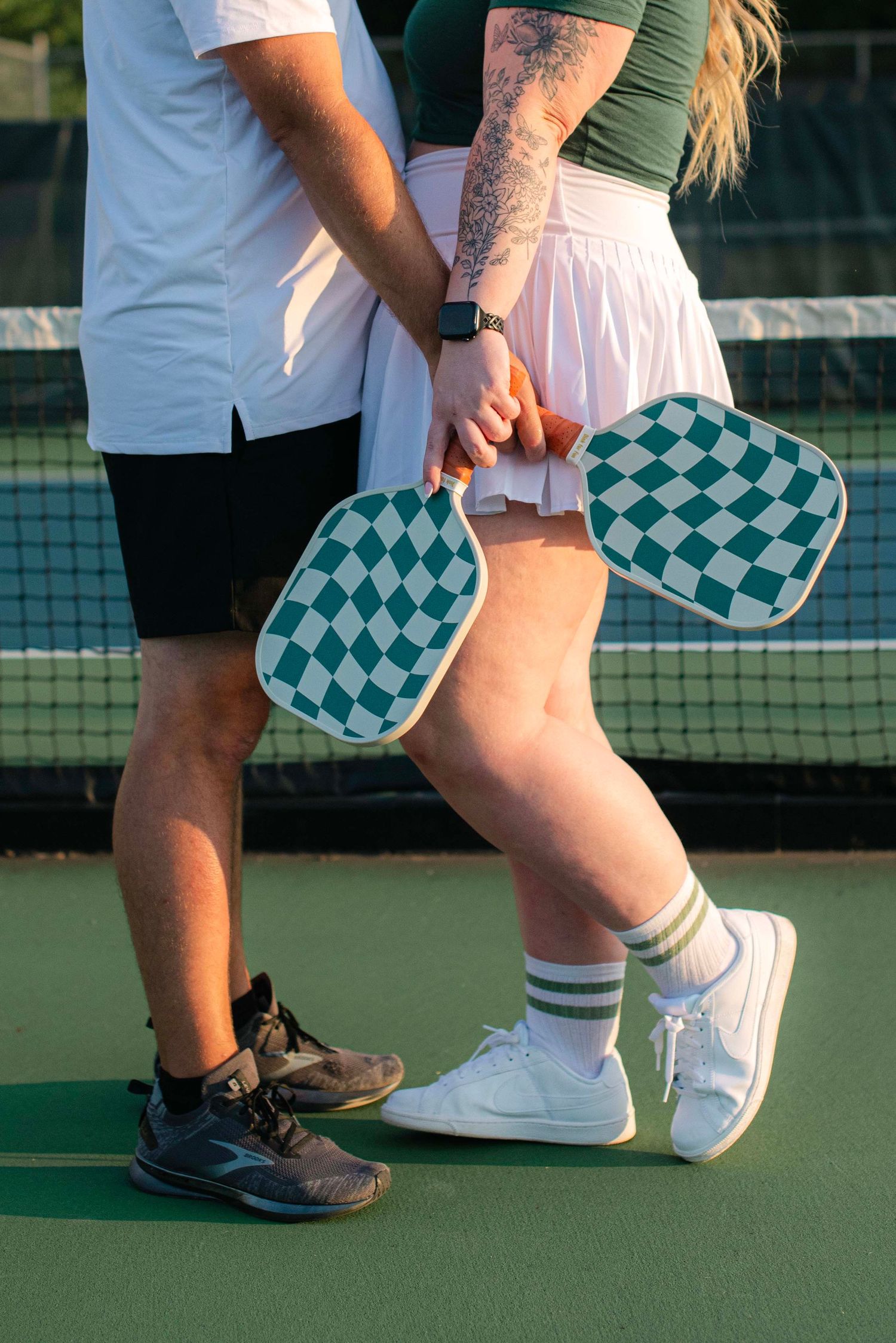 Two people wearing contrasting white and dark shoes stand close together on a green tennis court with checkered accessories.