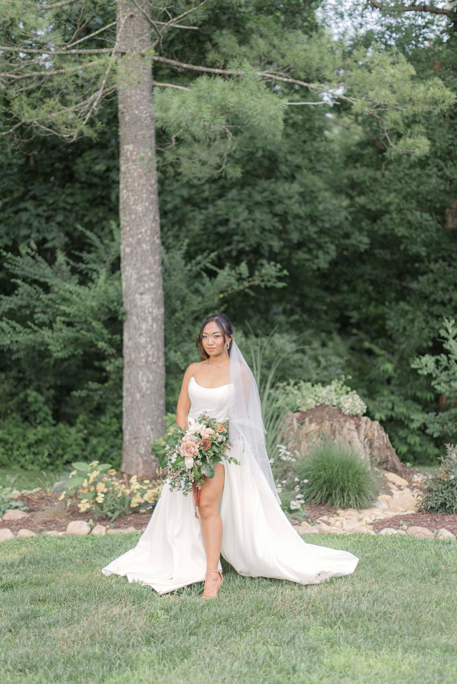 A bride in a flowing white wedding dress poses gracefully on a grassy lawn surrounded by lush green trees and landscaping.