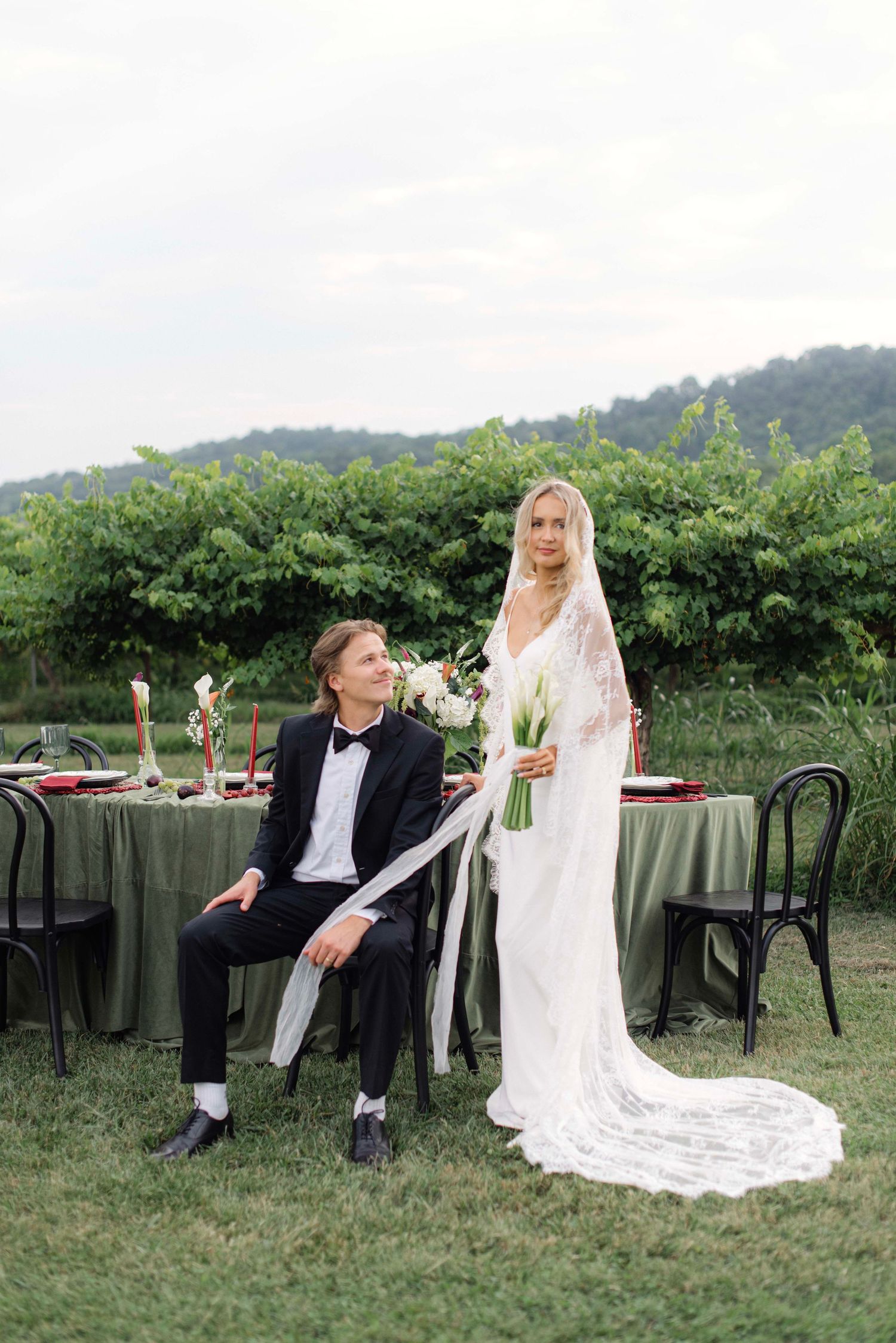 Wedding couple poses playfully on vintage chairs in an outdoor garden setting.